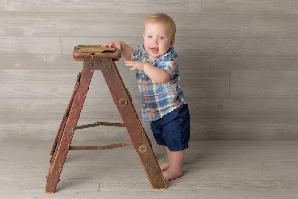Baby in plaid shirt stands beside vintage wooden ladder against gray background.