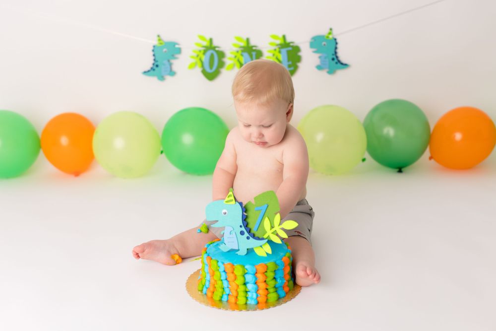 Baby sitting with blue and yellow birthday cake surrounded by green and orange balloons.