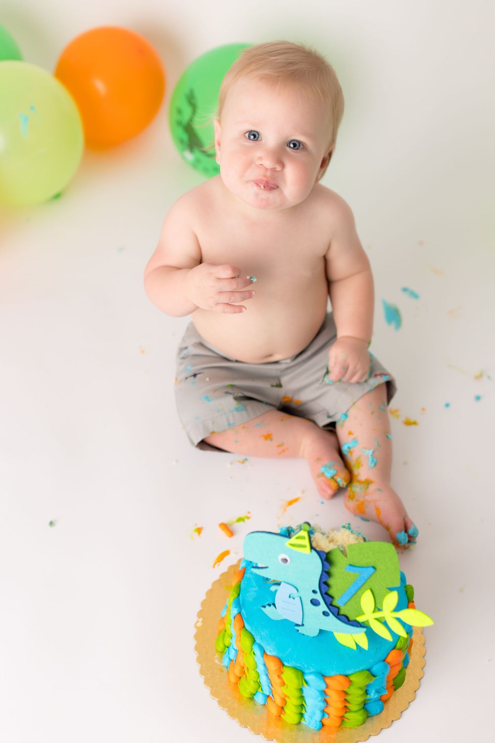 Messy baby with smeared cake sits among balloons with dinosaur-themed birthday decorations.