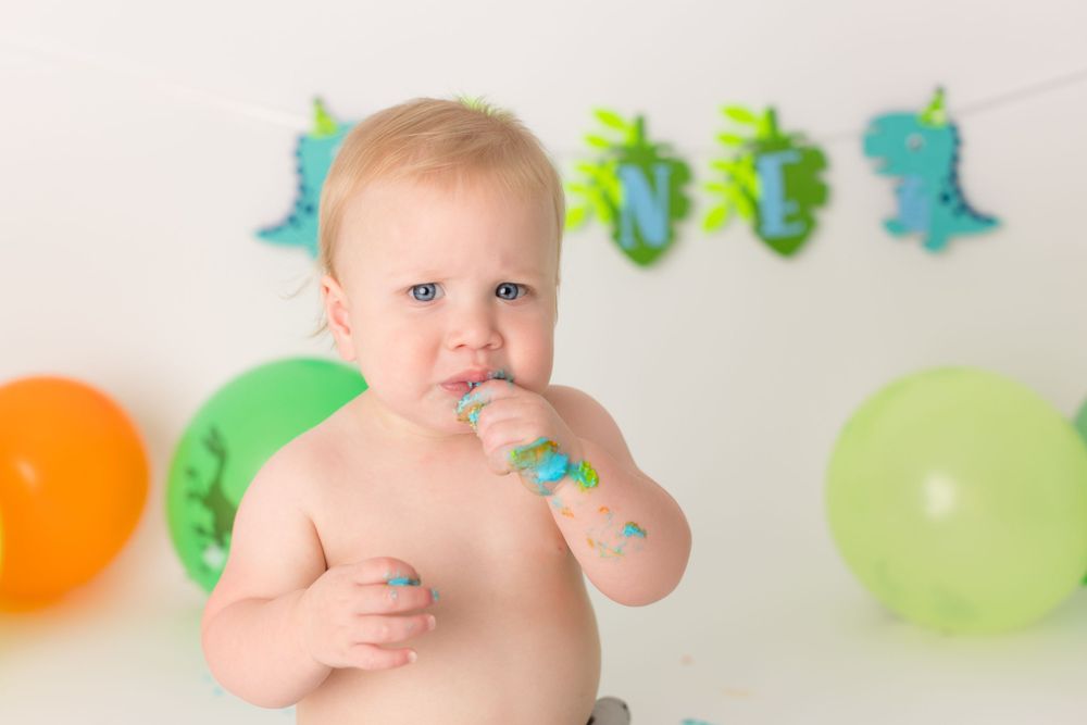 Baby with cake frosting on face sits among colorful balloons and dinosaur decorations.