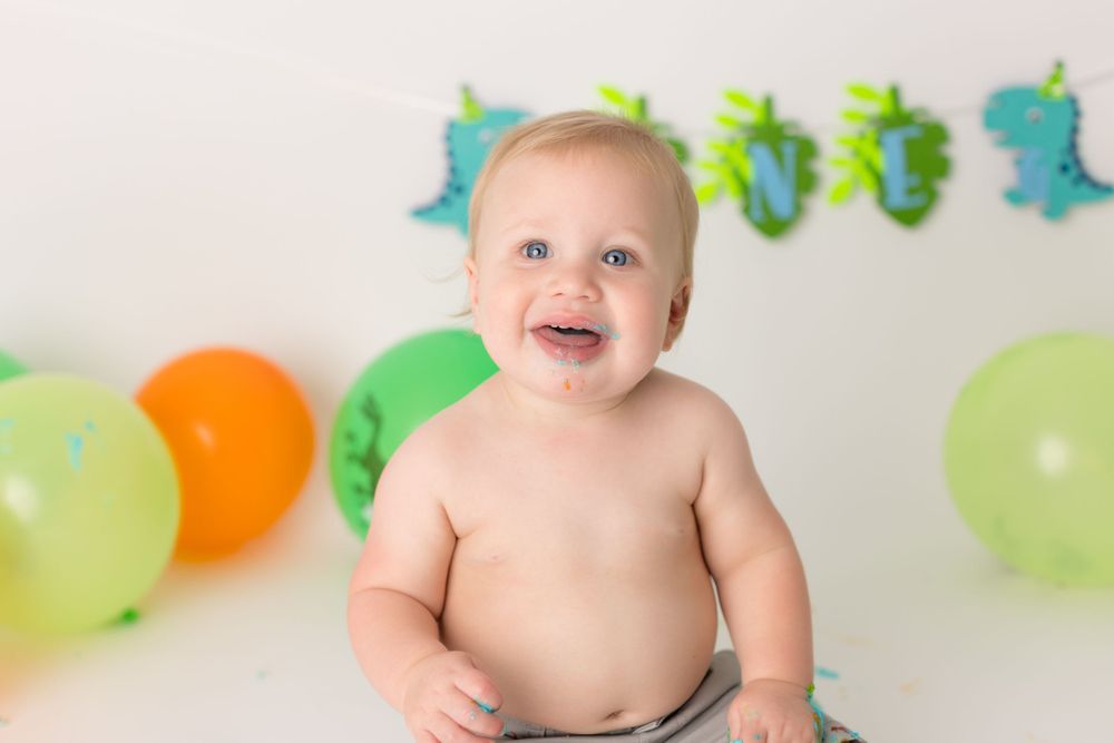 Smiling baby with dinosaur decorations and balloons during cake smash photo session.