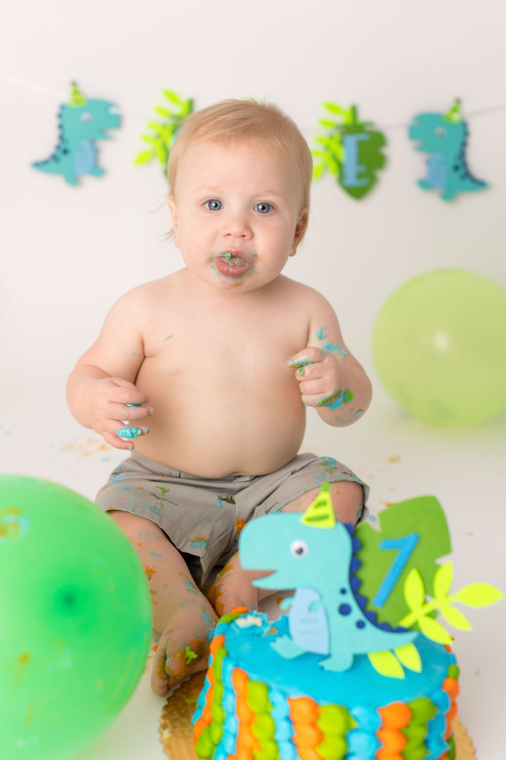 Baby with messy face from cake sitting among dinosaur decorations and balloons.