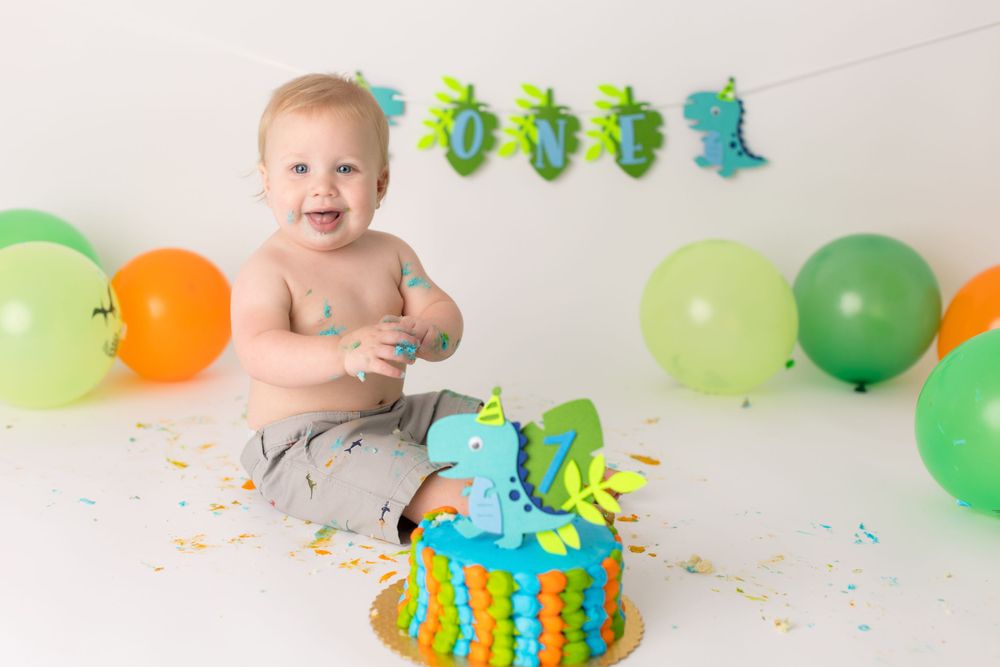 Baby celebrating first birthday with cake and balloons in green and orange party setup against white background.