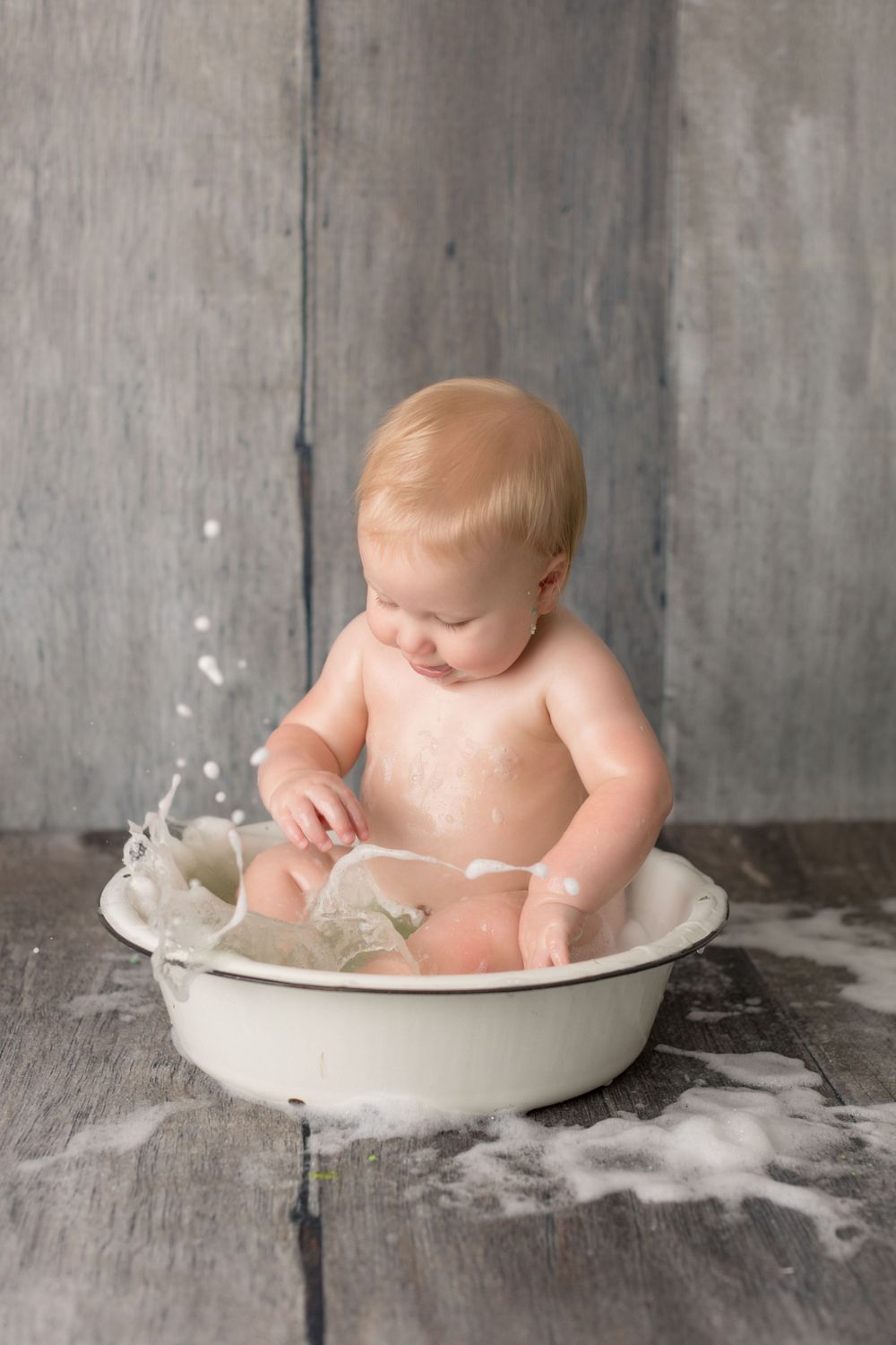 Baby sitting in white vintage wash basin with splashing milk against gray backdrop.