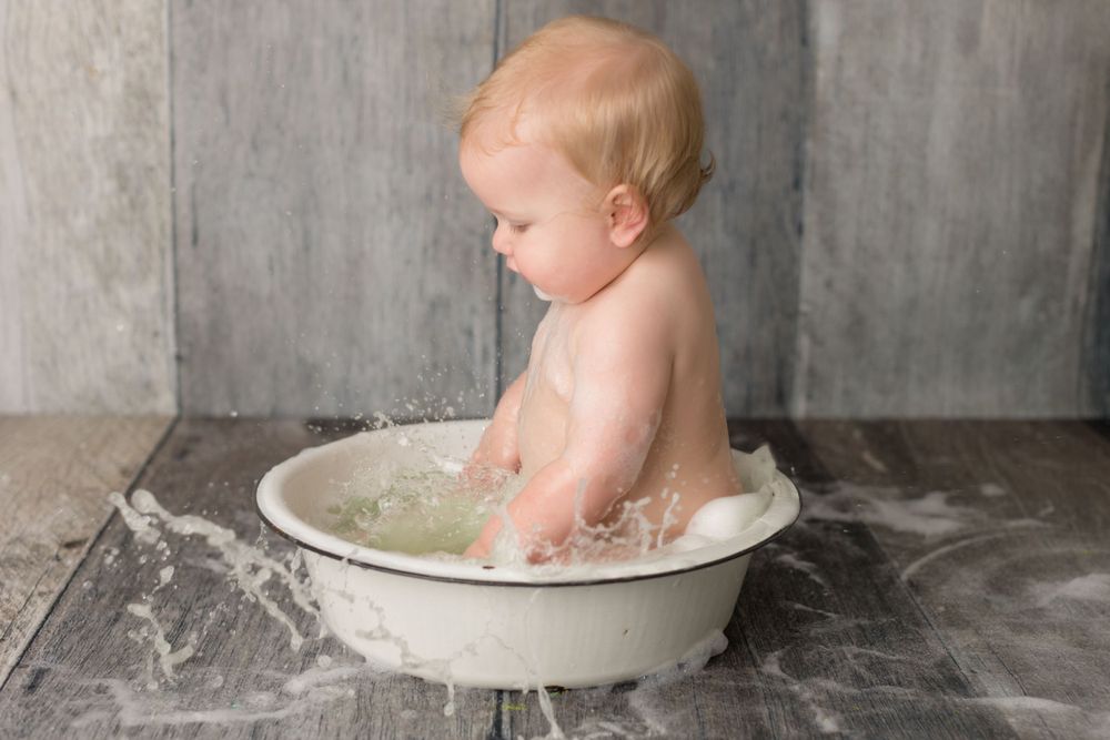 Baby playing in white vintage wash basin with milk splashes on gray wooden floor.