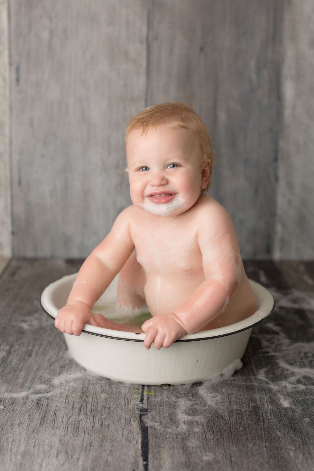 Baby splashing in vintage white basin against rustic gray backdrop.