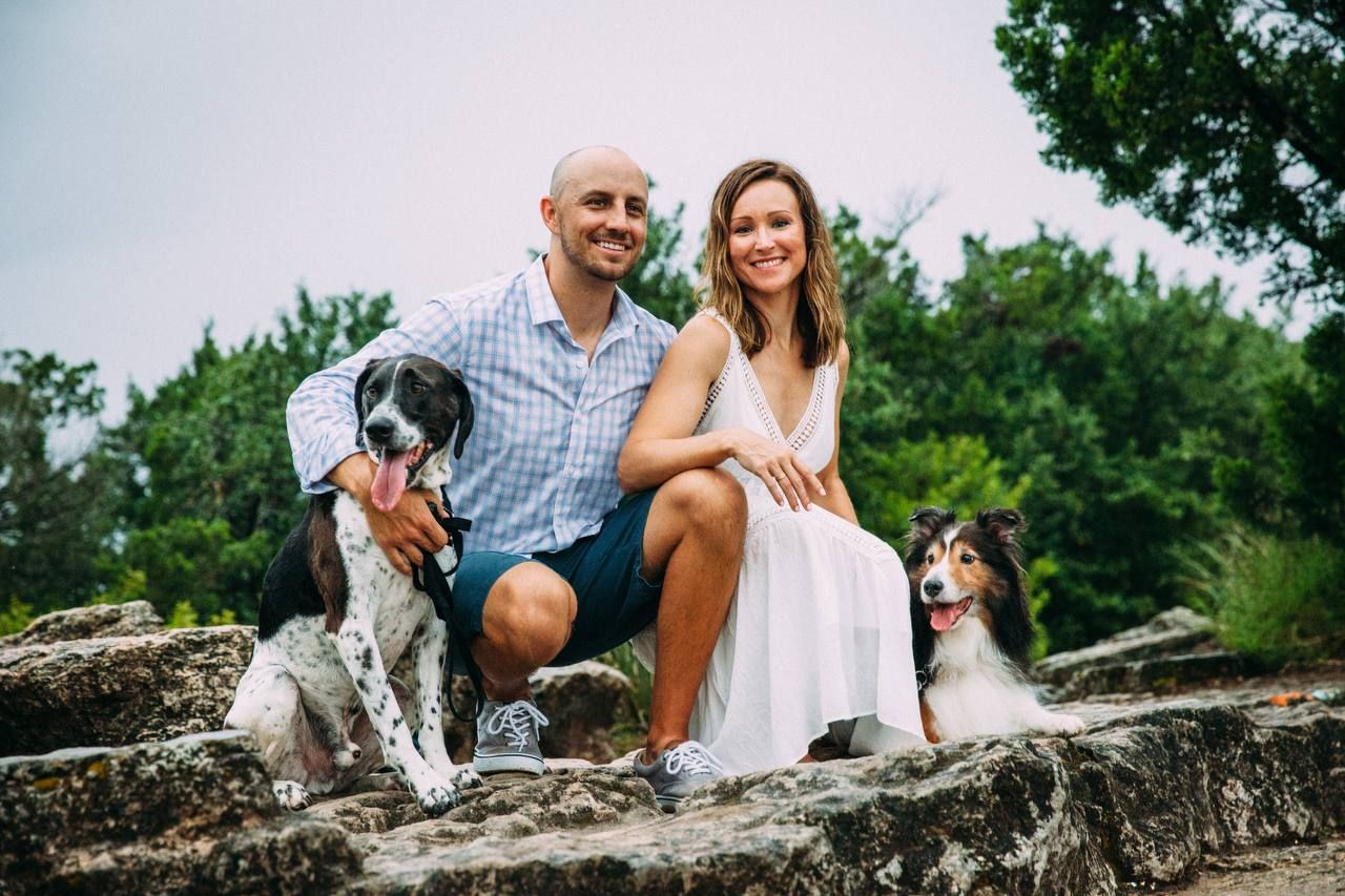 A couple sits on stone steps with their two dogs against a nature backdrop.