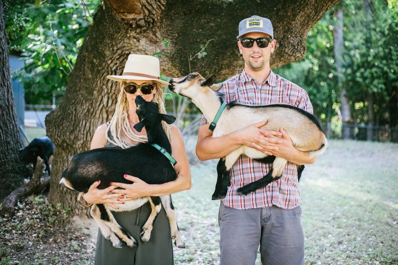 Two people in casual attire holding their dogs while standing beneath a large tree.