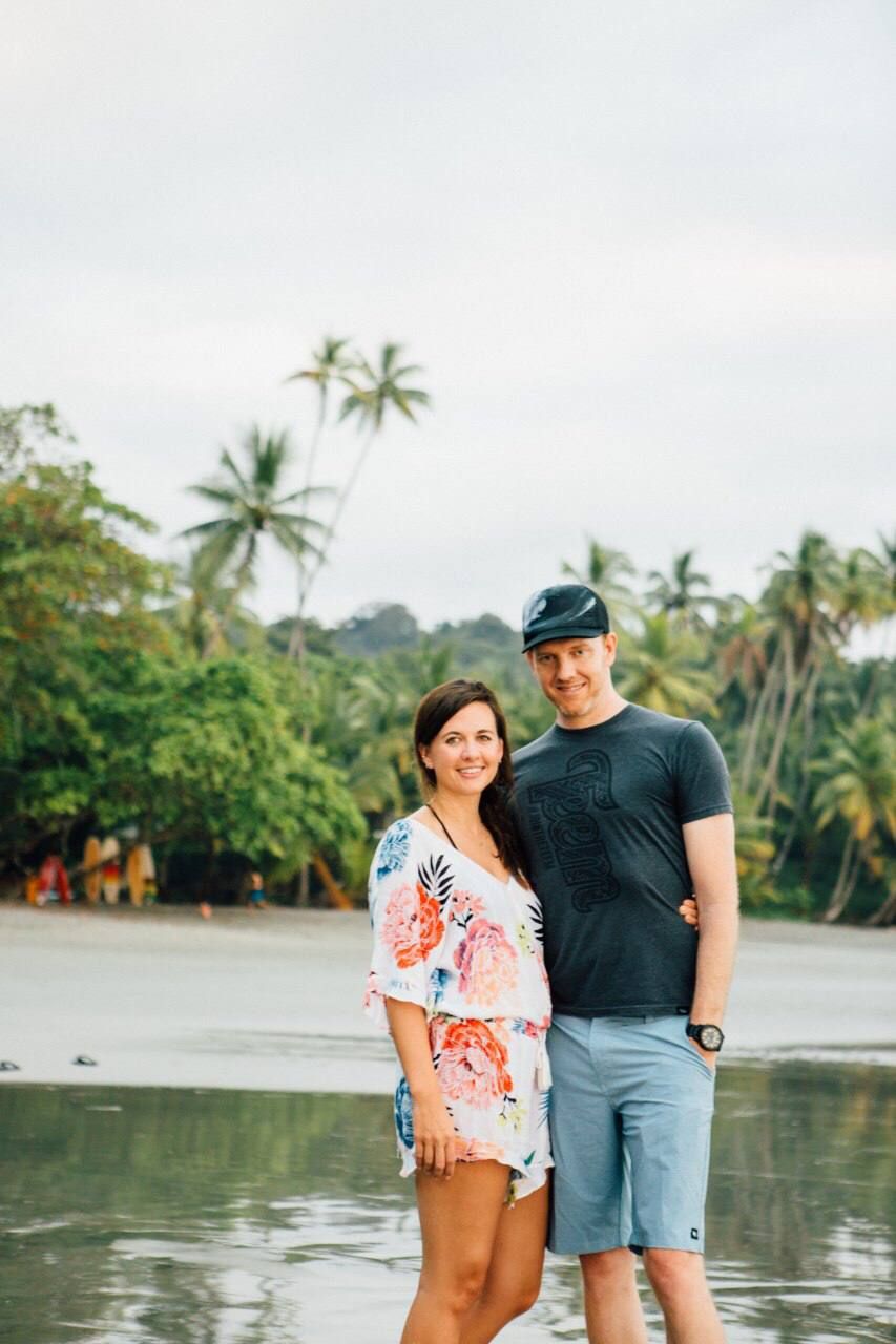 A couple poses together on a tropical beach with palm trees and cloudy skies in the background.