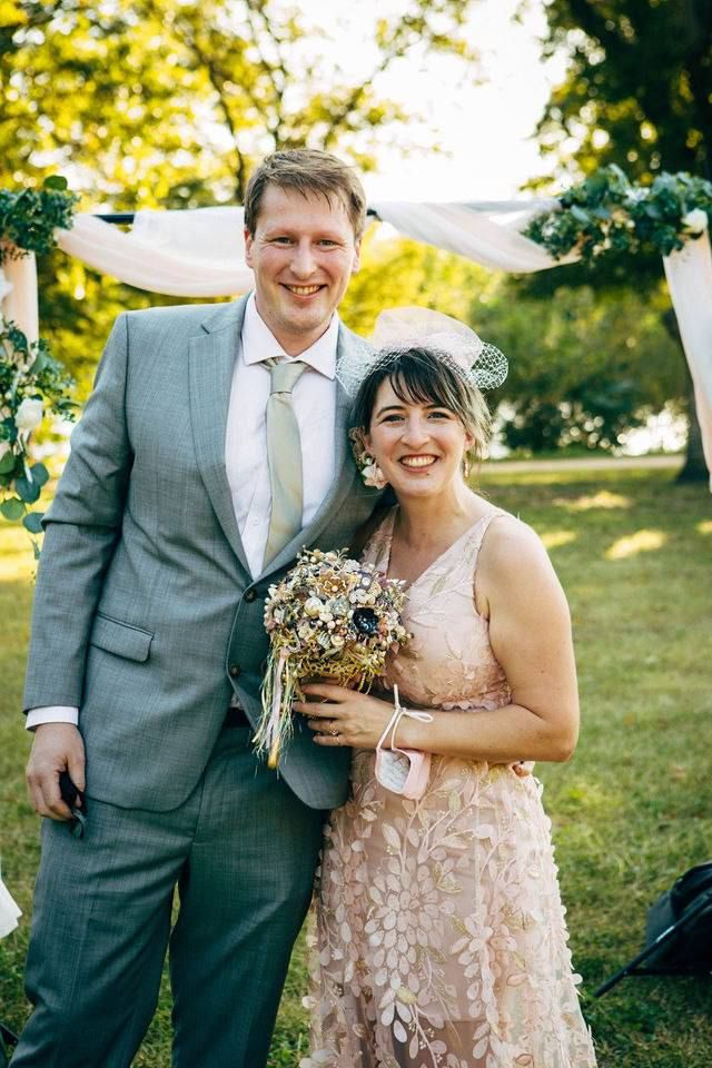 A wedding couple in formal attire pose together under an outdoor ceremony arch with flowers.