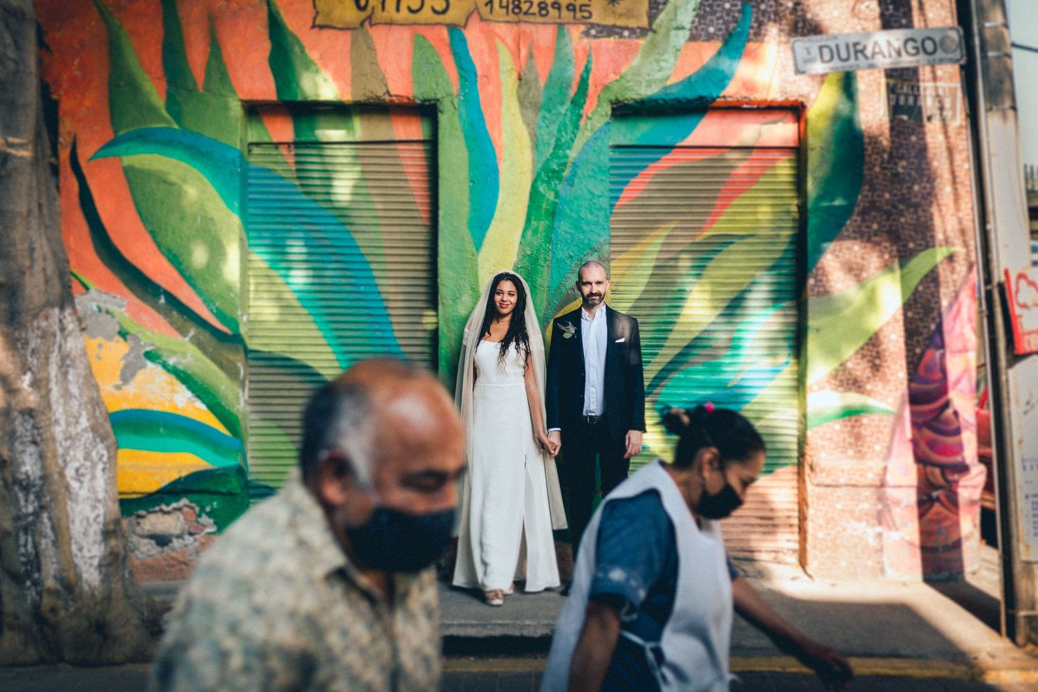 Wedding couple standing in front of vibrant green street art while pedestrian walks past.