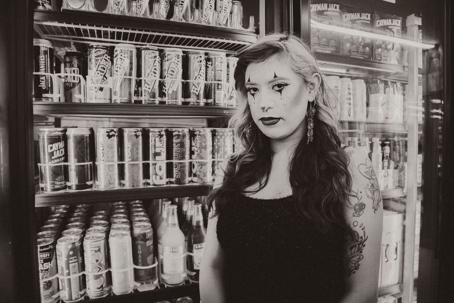 Black and white photo of figure in dark attire standing against shelves of bottles.