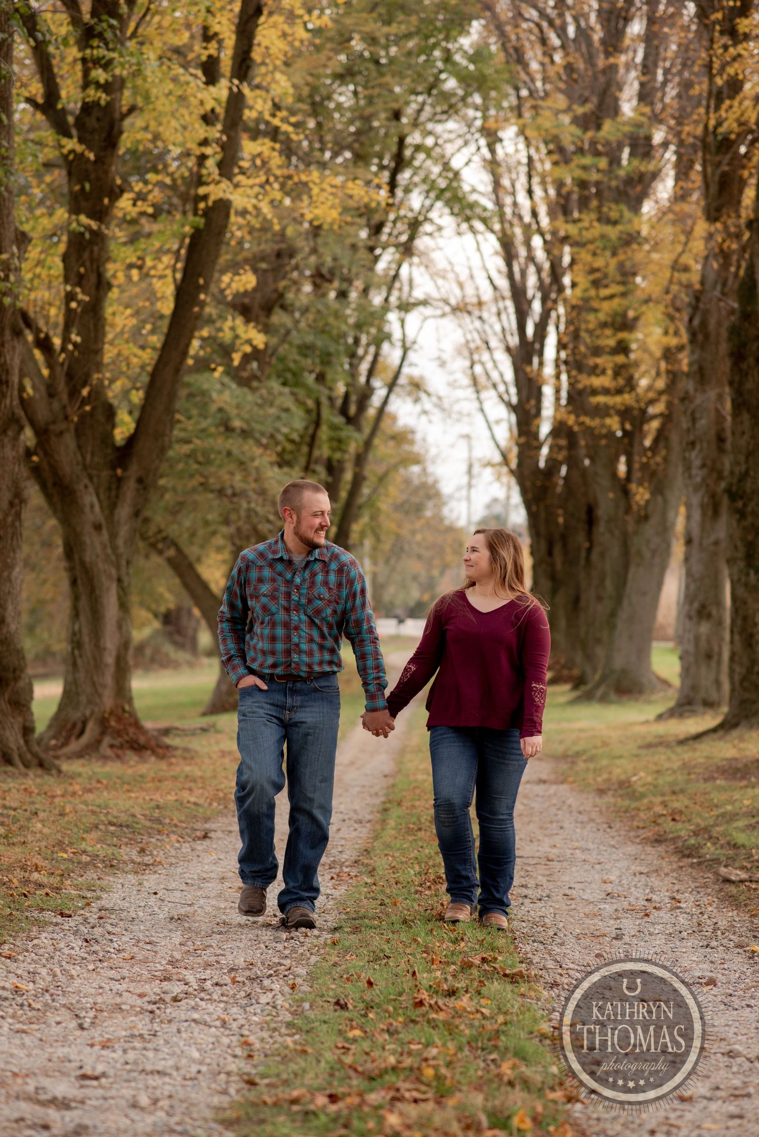 A couple walks hand in hand down a tree-lined path covered in fallen autumn leaves.