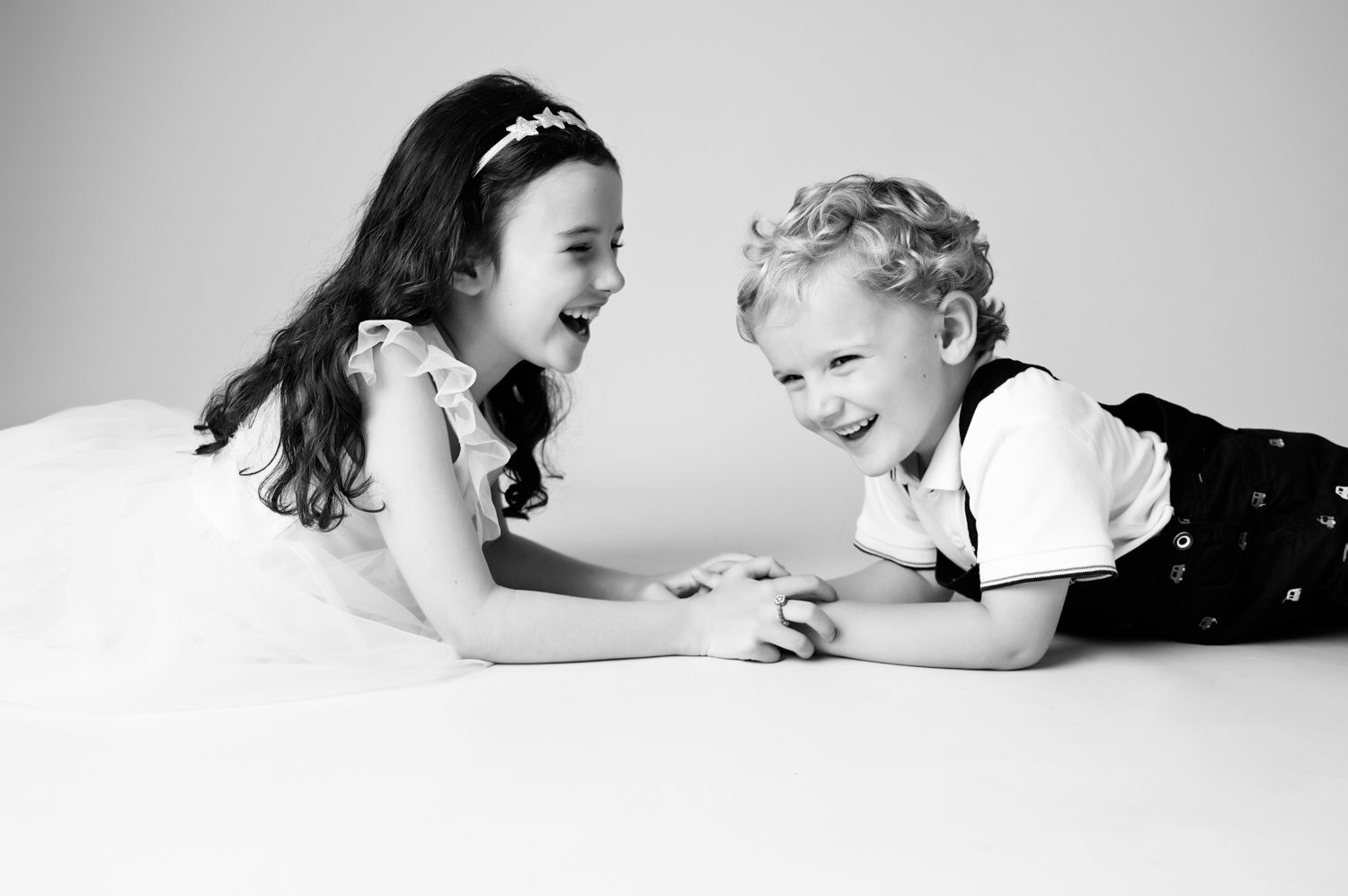 Two children laying on white background laughing and playing together in black and white photograph.