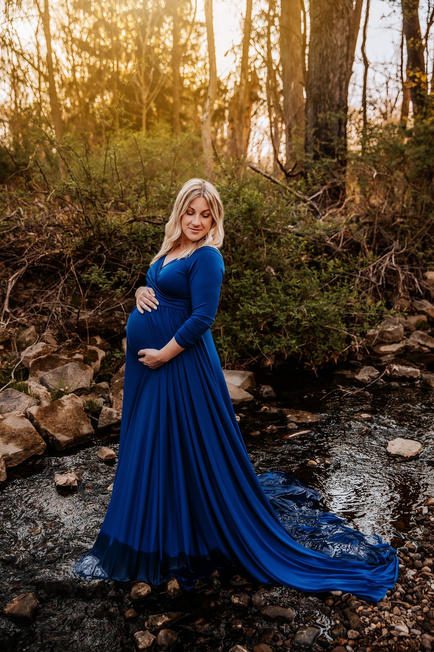 Maternity photo of woman in flowing blue dress by stream at sunset.