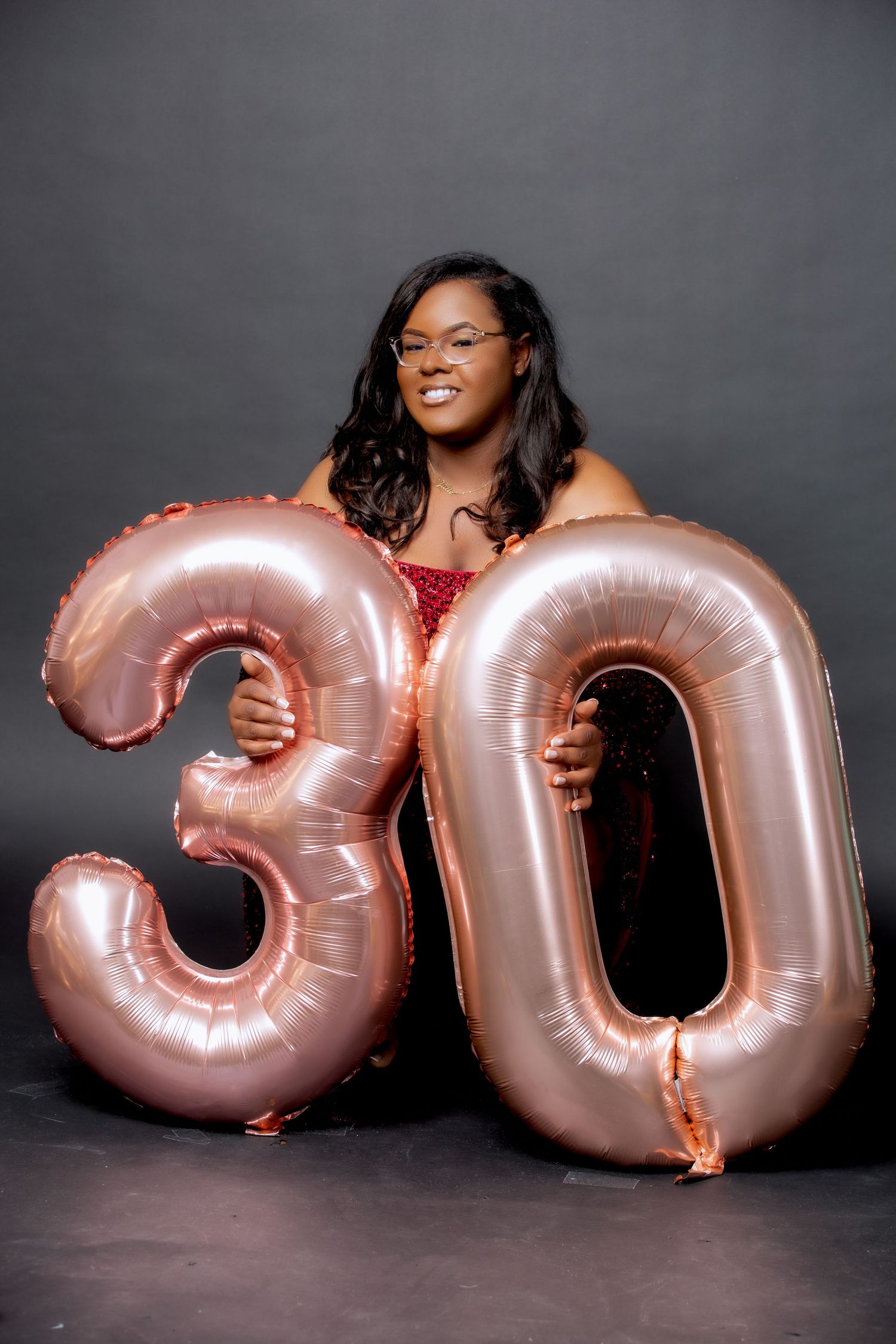 Woman in sparkly dress posing with large rose gold number 30 balloons against dark background.