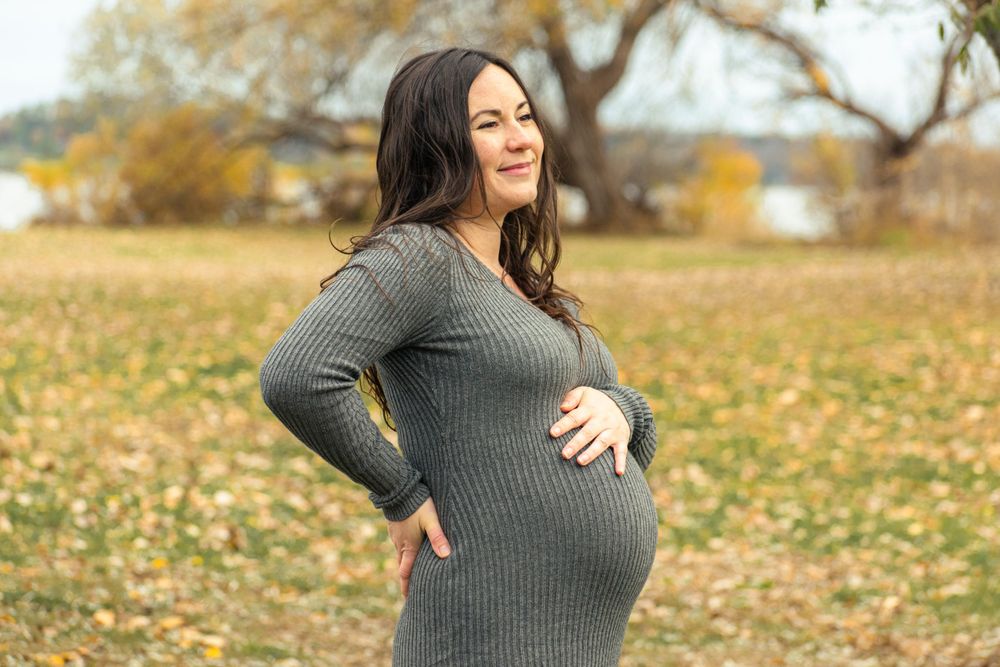 Maternity photo showing pregnant figure in grey sweater dress with fall foliage in background.