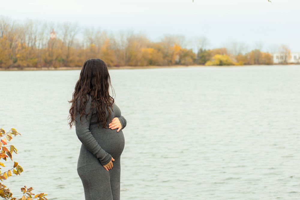 Pregnant silhouette in grey dress standing by a lake with autumn trees in background.