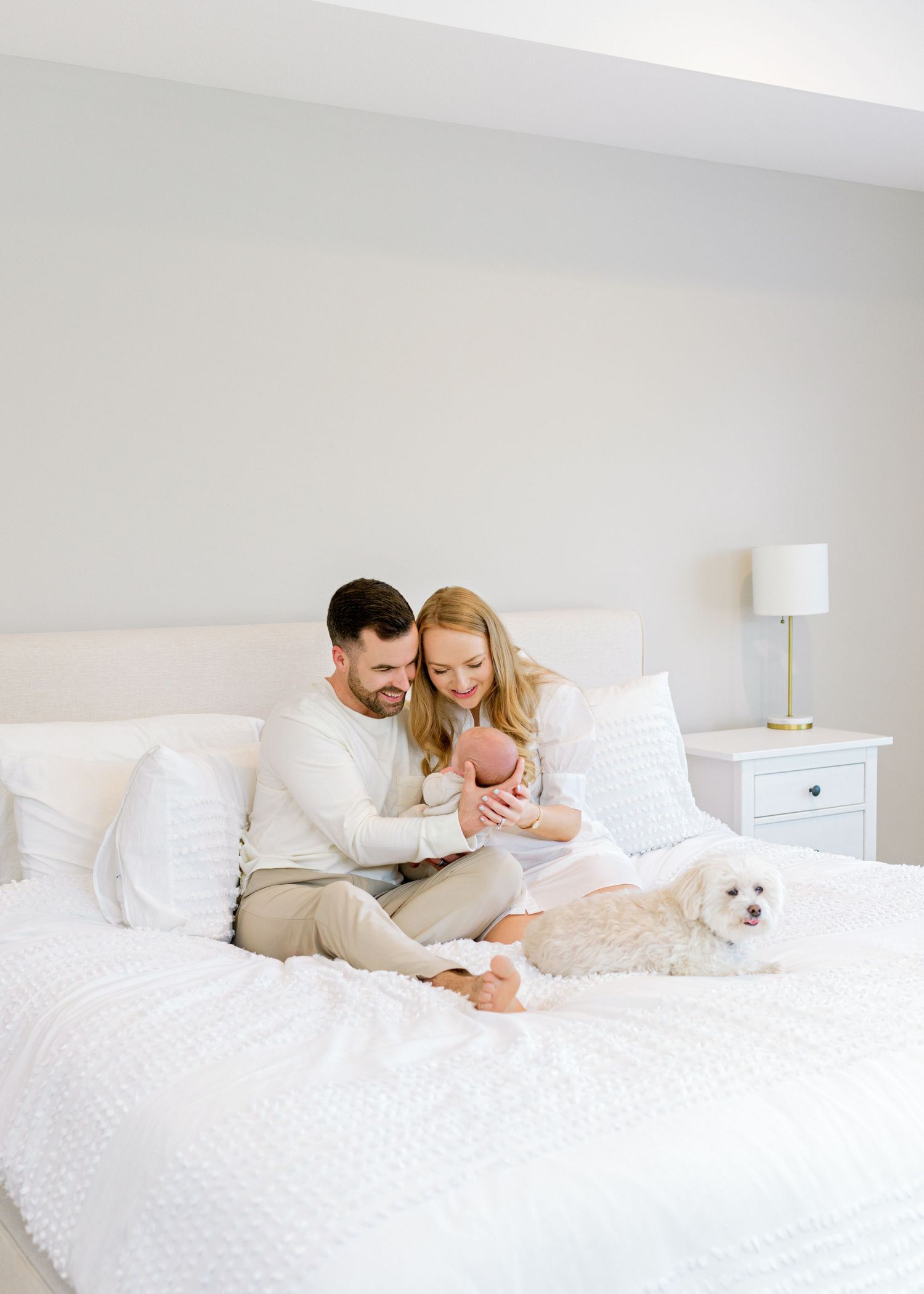 austin, tx parents sitting on their bed, holding their newborn son as they look down at him smiling.