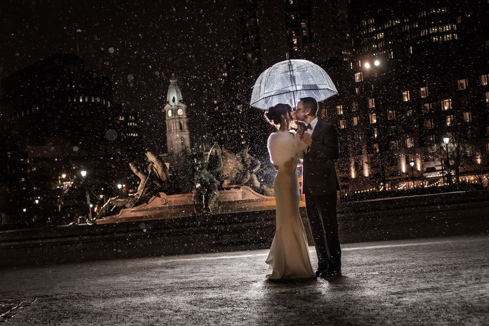 Couple shares romantic moment under clear umbrella at night with city fountain in background.