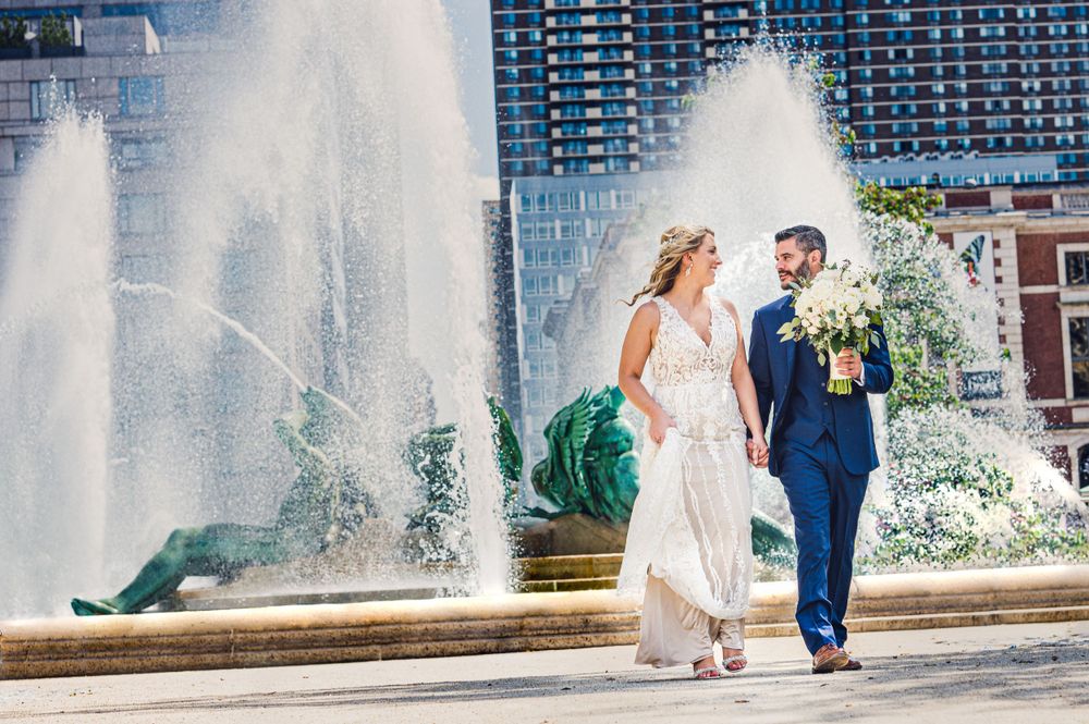 Couple in wedding attire walks past grand fountain with city buildings in background.