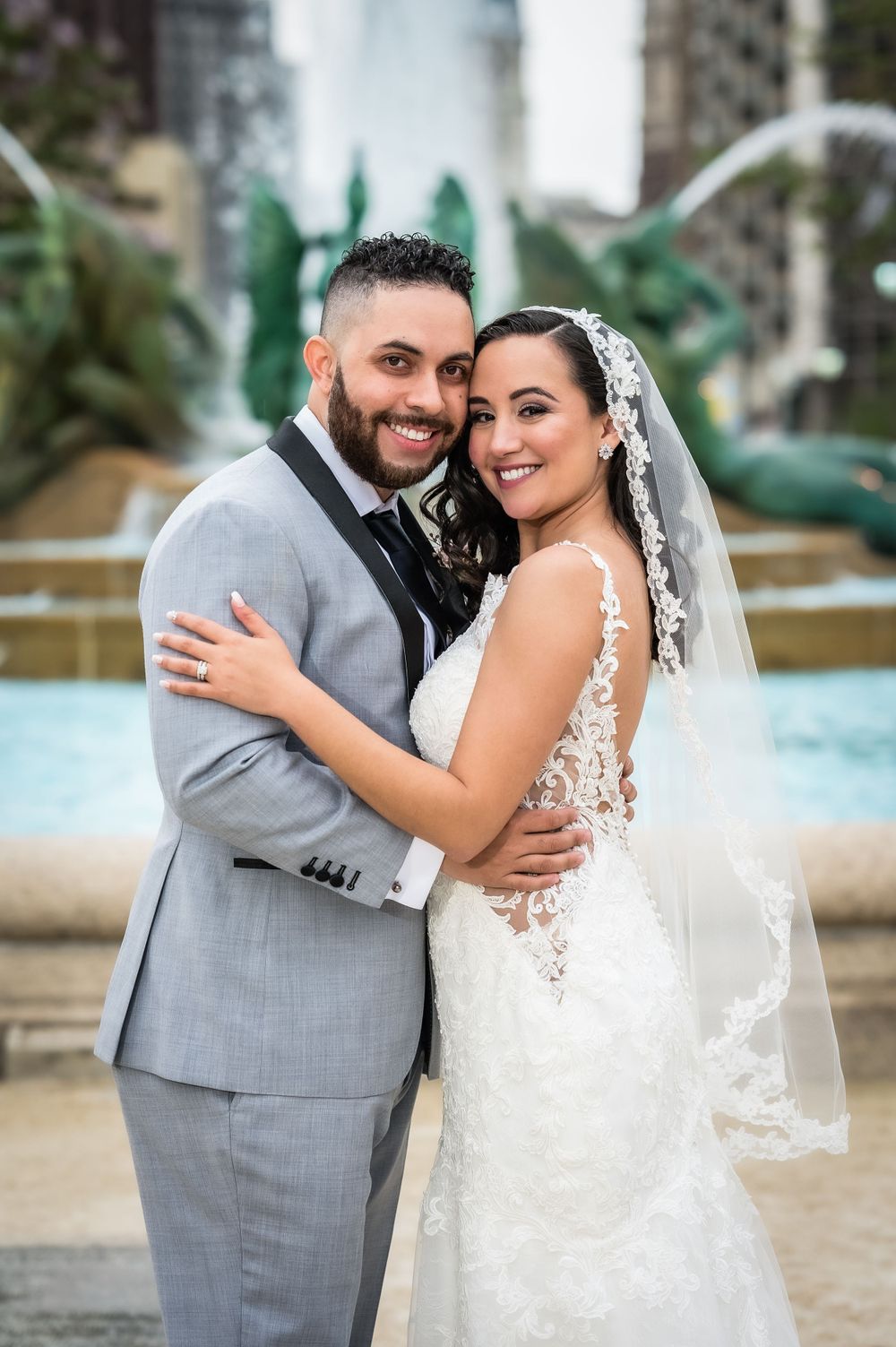 Couple in wedding attire poses together in front of ornate fountain with city backdrop.