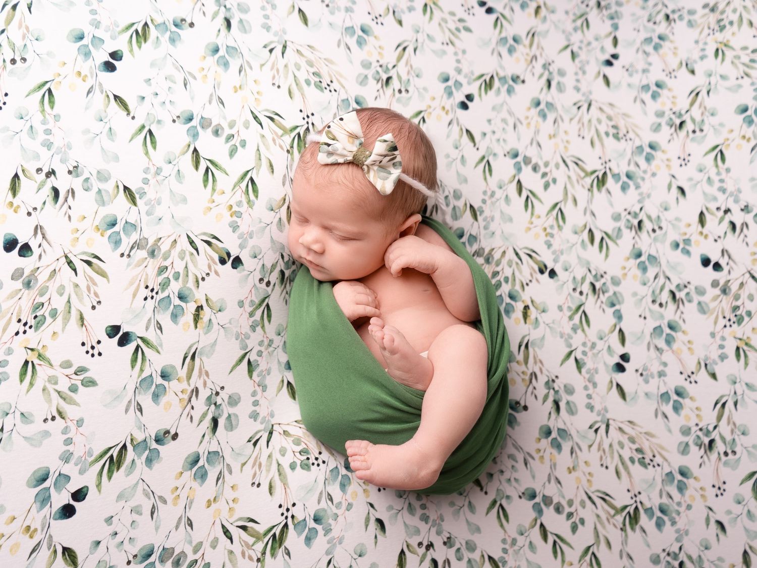 Baby girl sleeping on neutral eucalyptus backdrop, wrapped in green with bow headband at newborn photoshoot in Doncaster