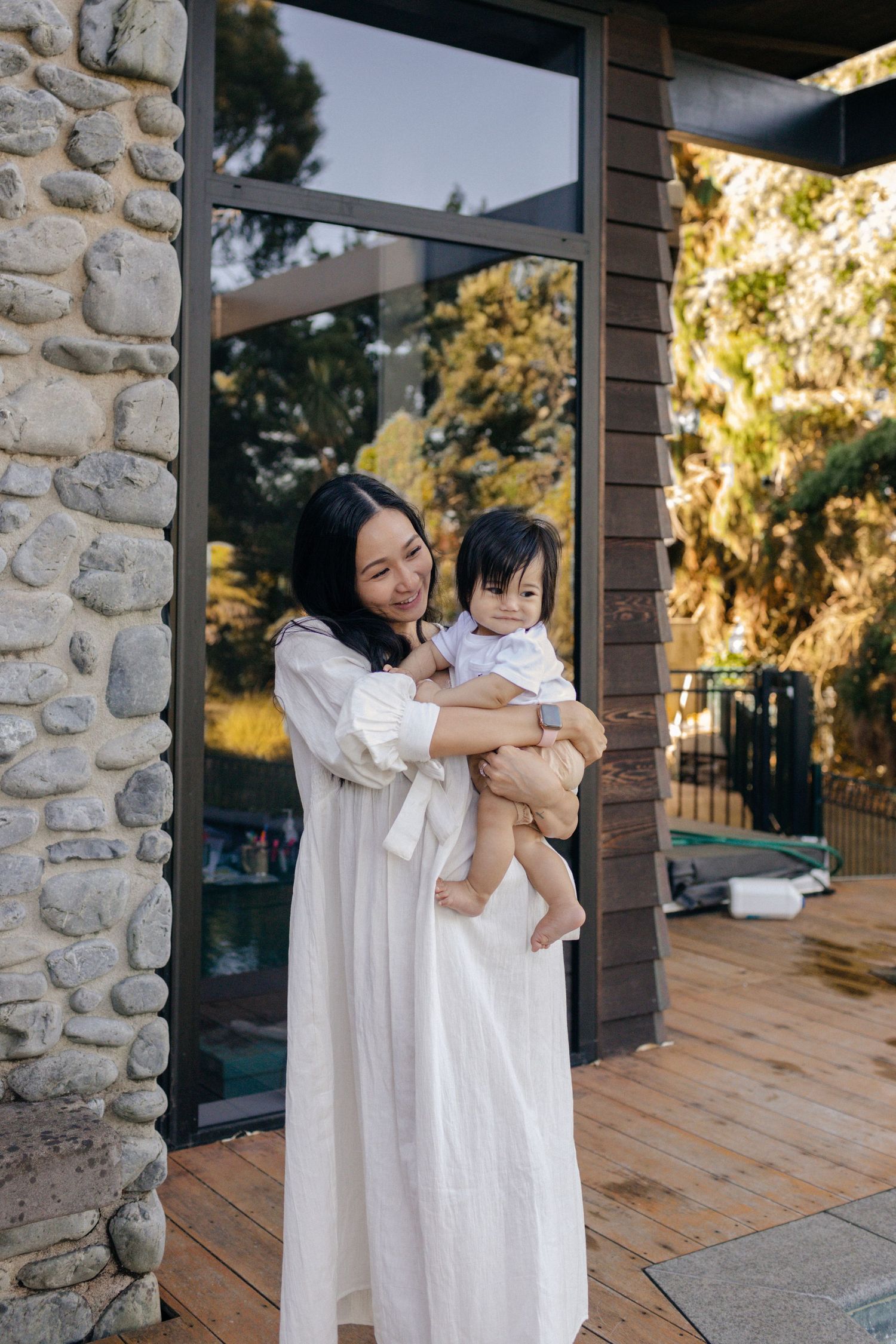 Mother and child in matching white dresses standing on wooden deck by stone wall.