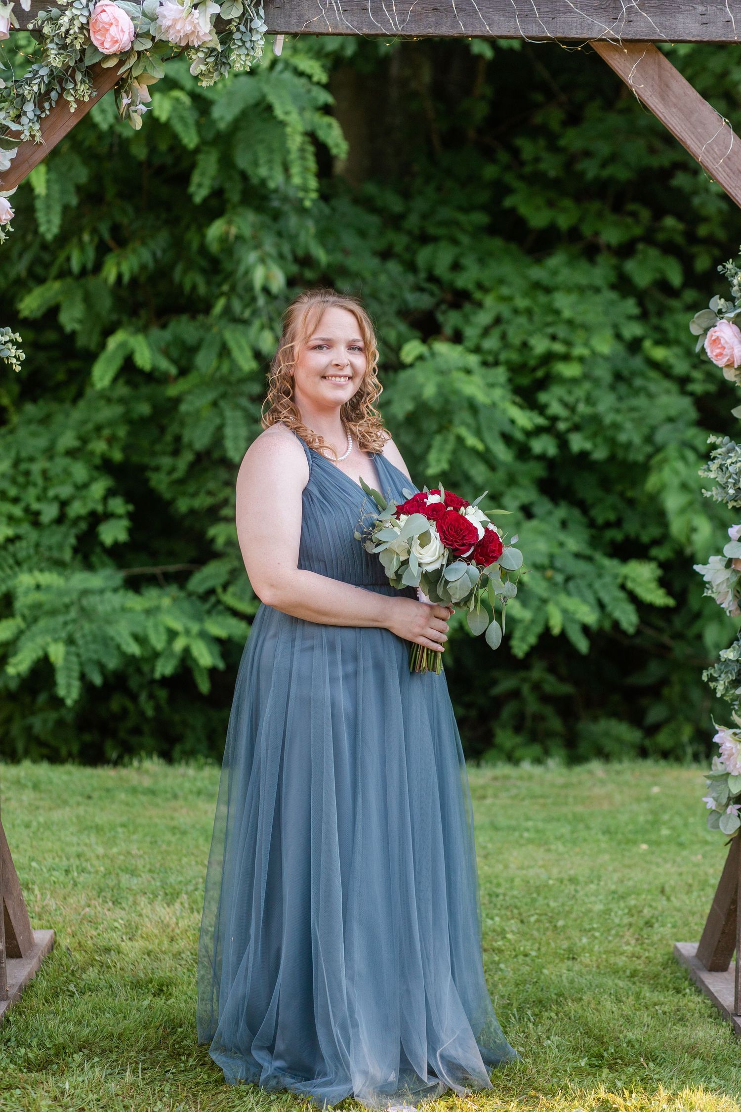Bridesmaid in flowing dusty blue dress holding red and white rose bouquet in garden setting.