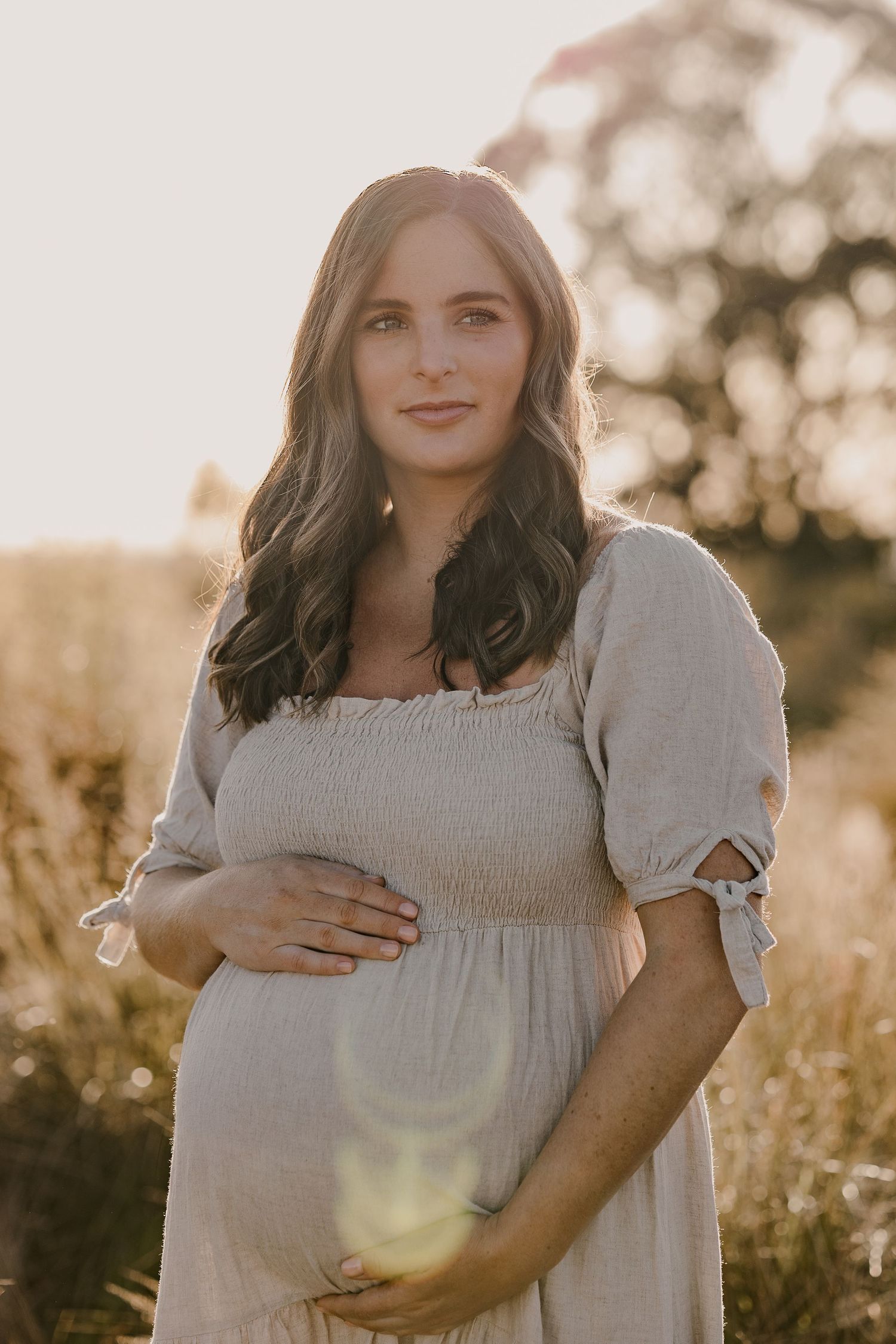 Expectant mother in white dress stands in golden sunlit field during sunset.