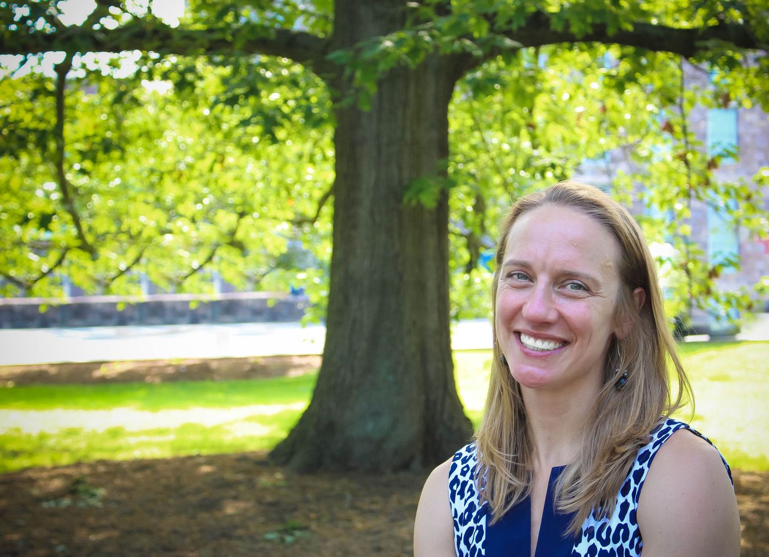 A person smiles warmly while standing in front of a large tree in a sunny park setting.