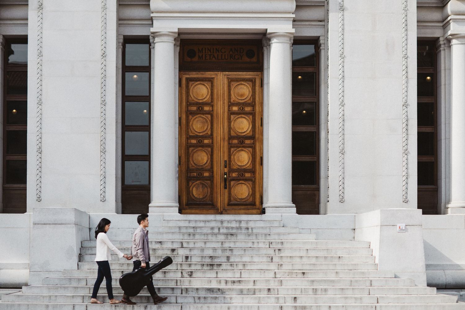 Two people walking up white marble steps toward large wooden doors of classical building.