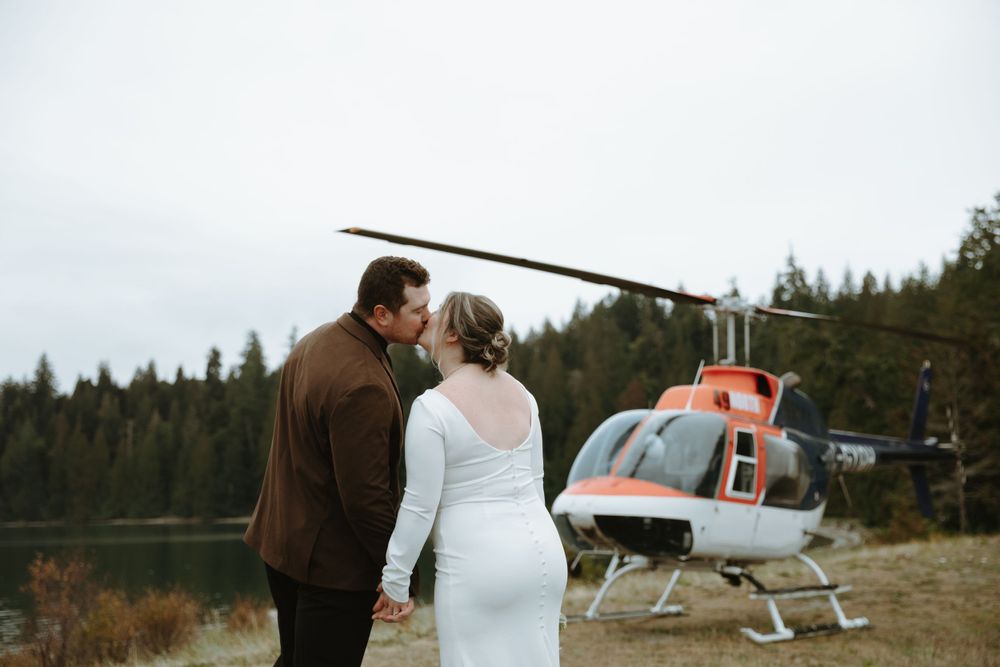A couple stands together looking at a helicopter in a mountainous forest landscape.