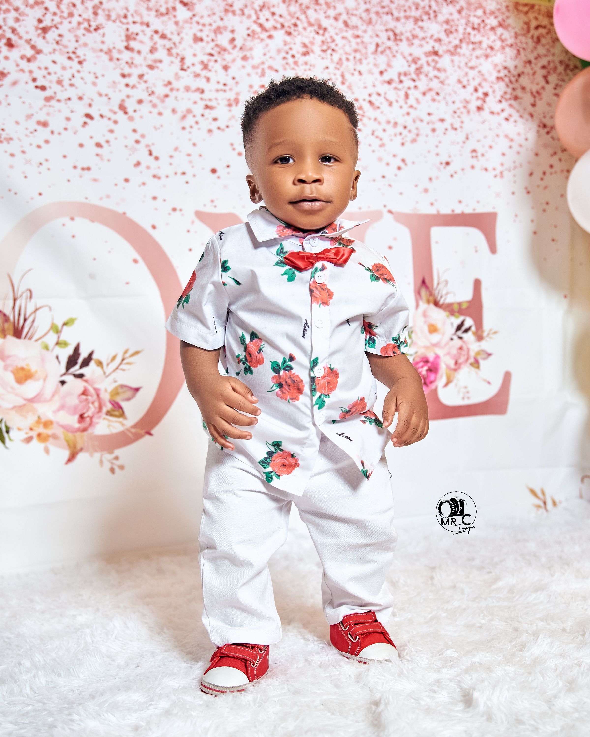 Child in white floral outfit with red bowtie poses against backdrop with pink confetti and letters.