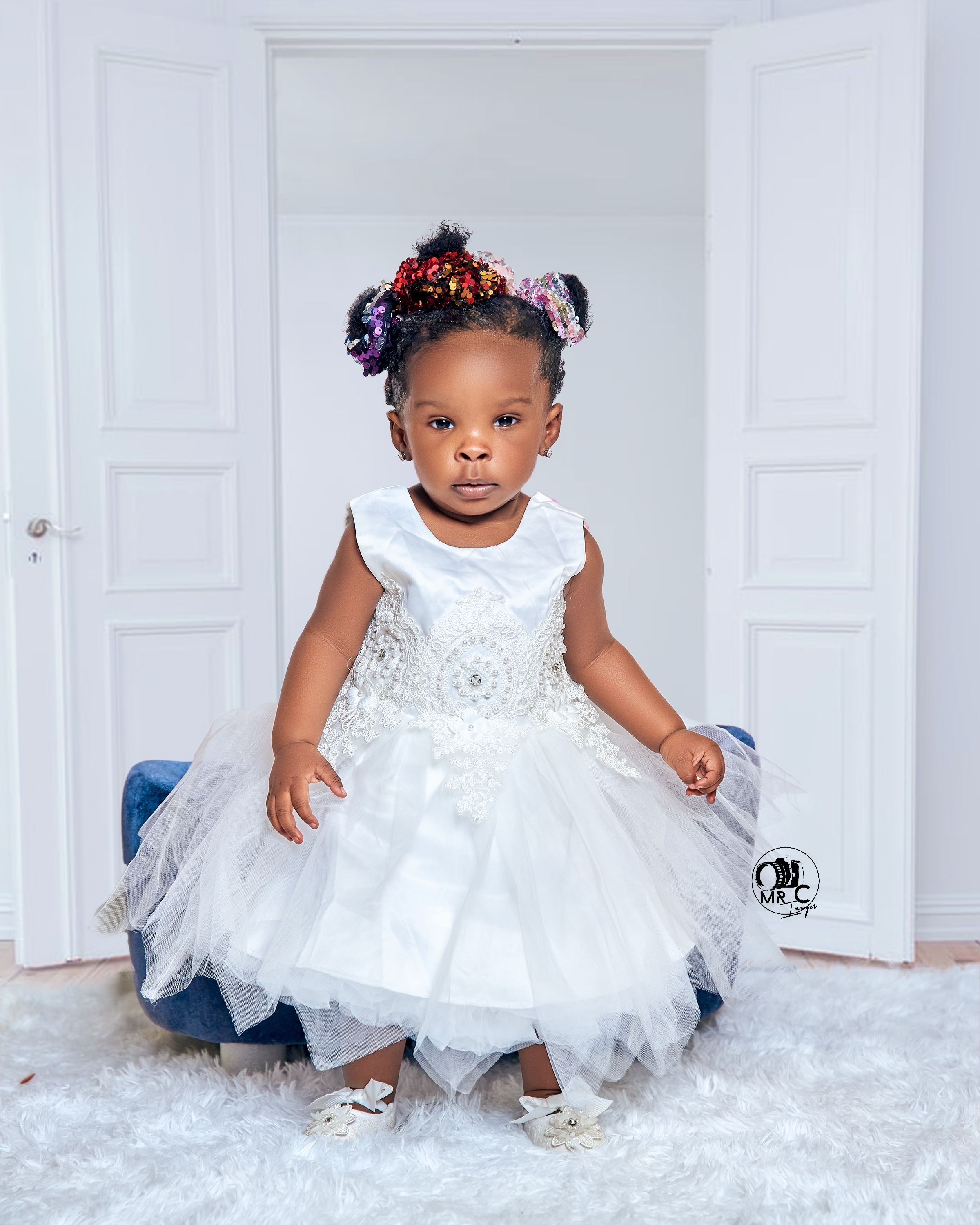Child in white tulle dress with sparkly details poses against white doorway backdrop.
