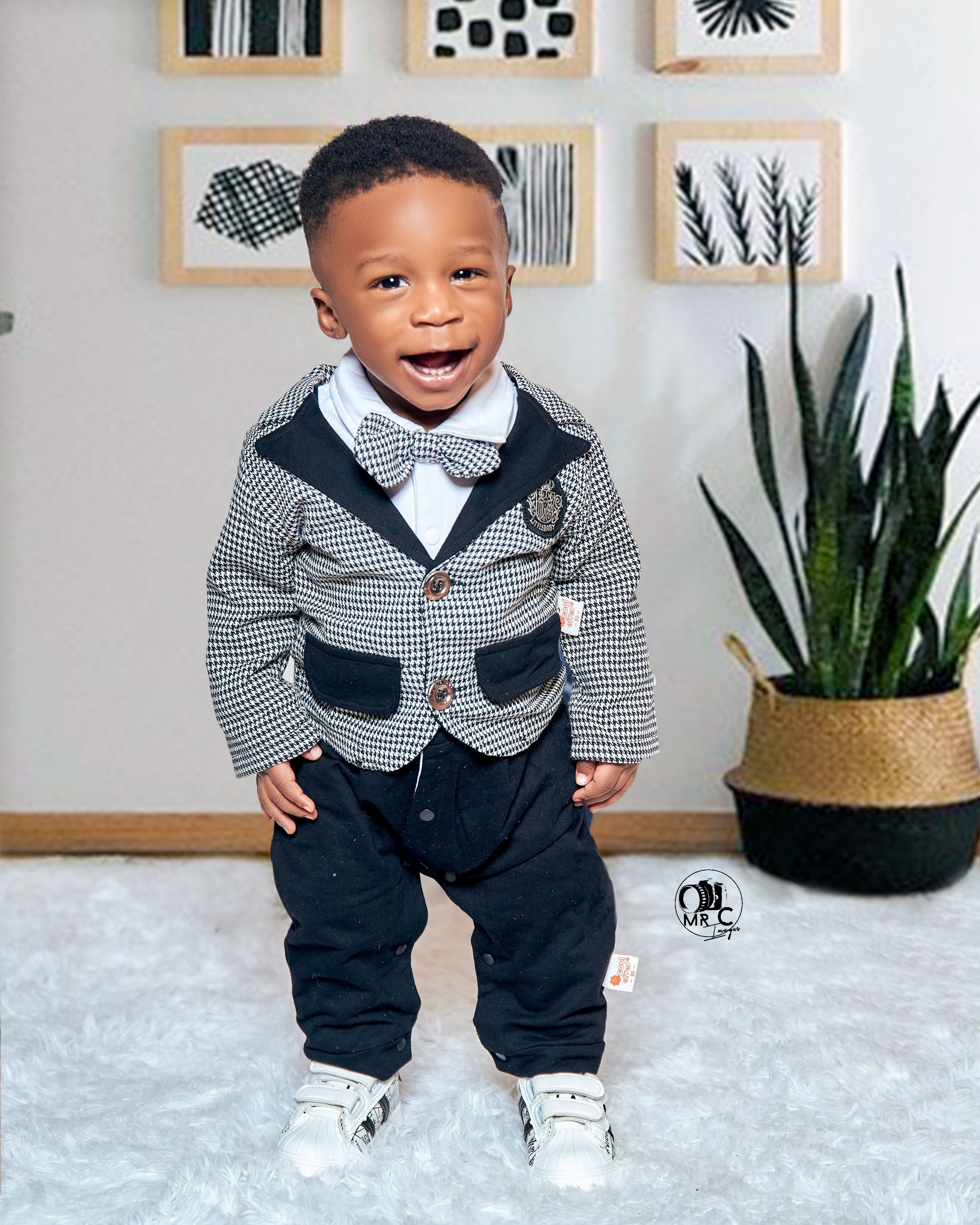 Child in formal houndstooth jacket and bowtie smiles against wall with framed artwork.