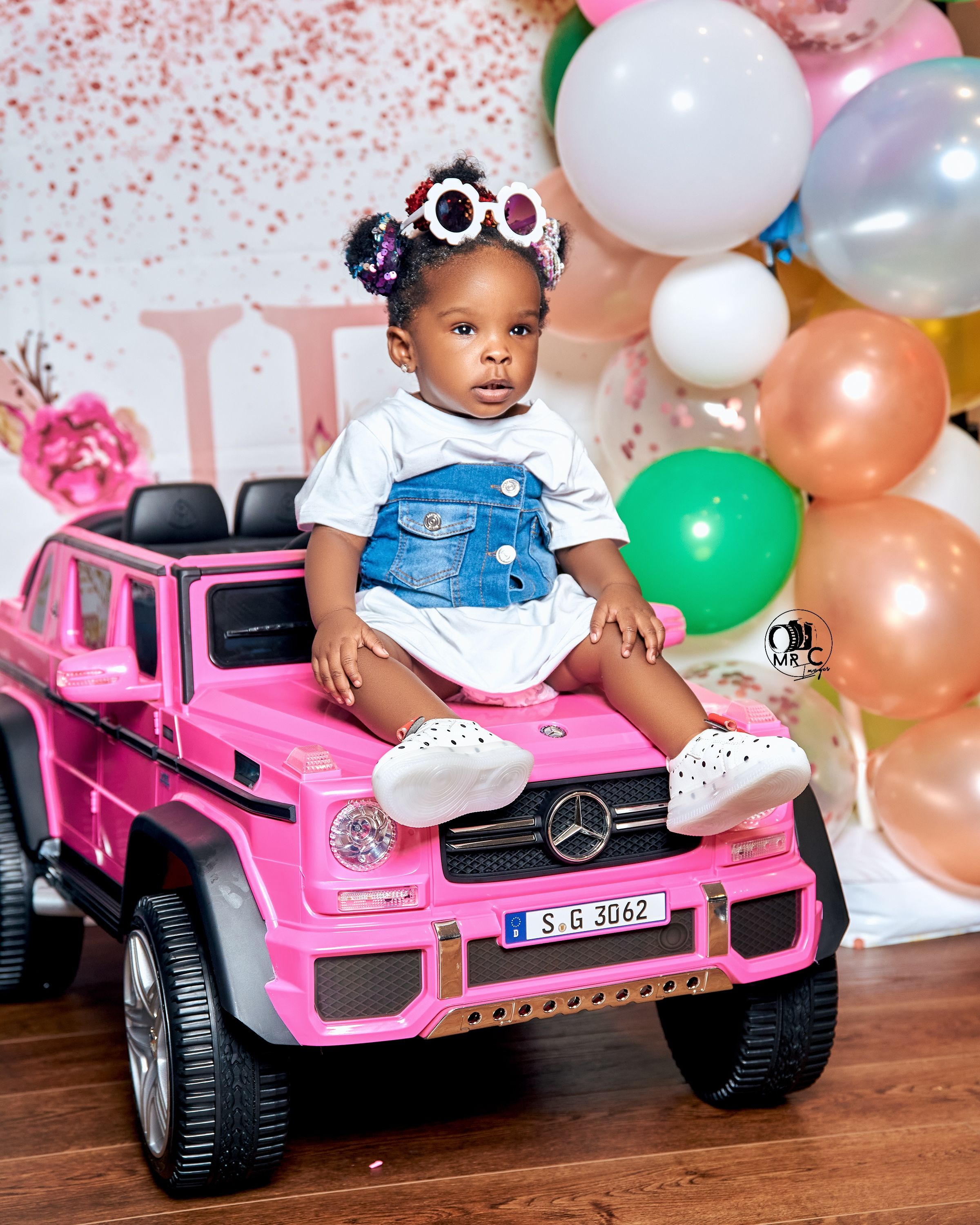 Child wearing denim overalls sits on pink toy Mercedes ride-on car surrounded by colorful balloons.