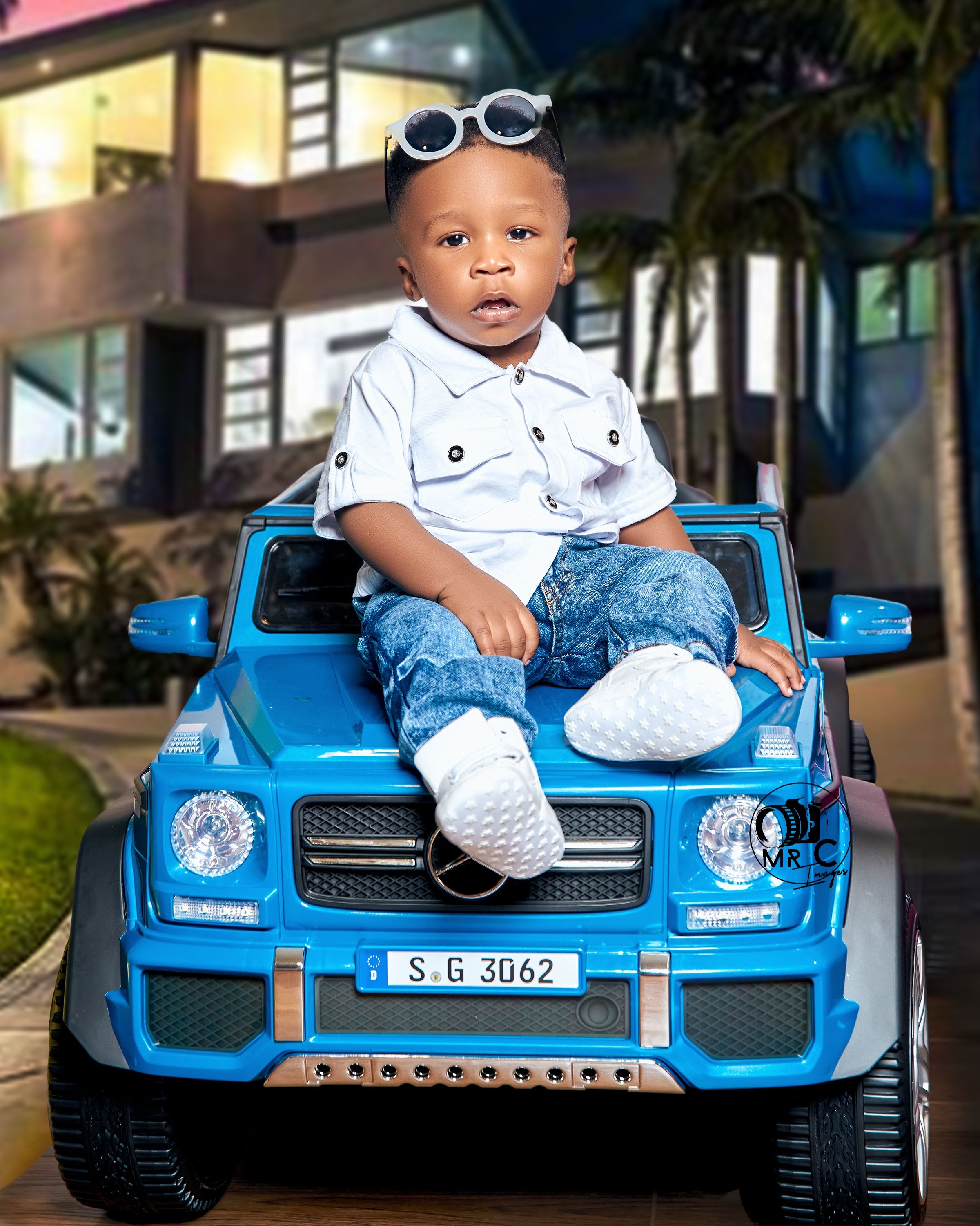 Child in casual outfit sits on bright blue toy Mercedes with sunglasses on head.
