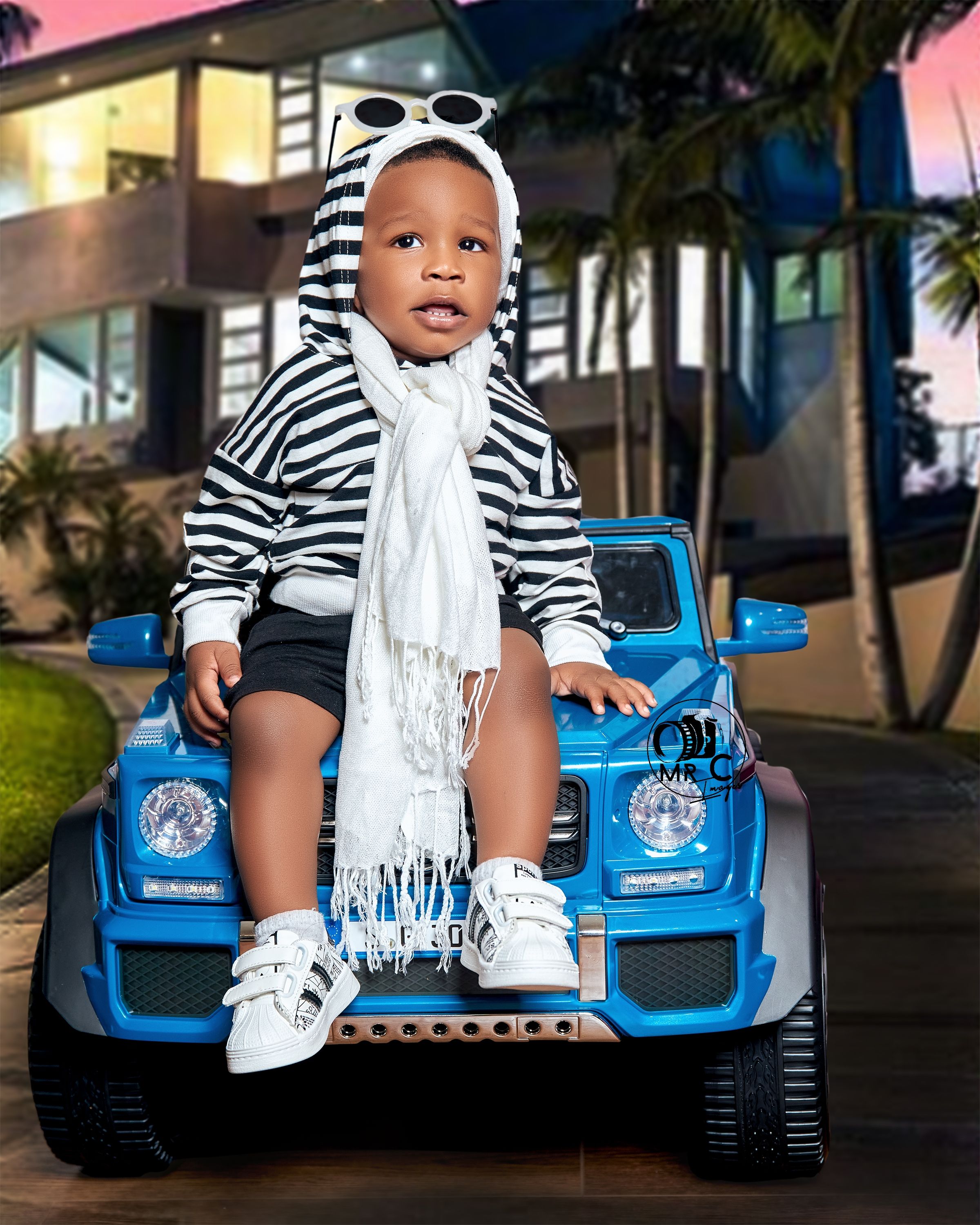 Child in zebra print outfit with white scarf poses on blue toy car outdoors.