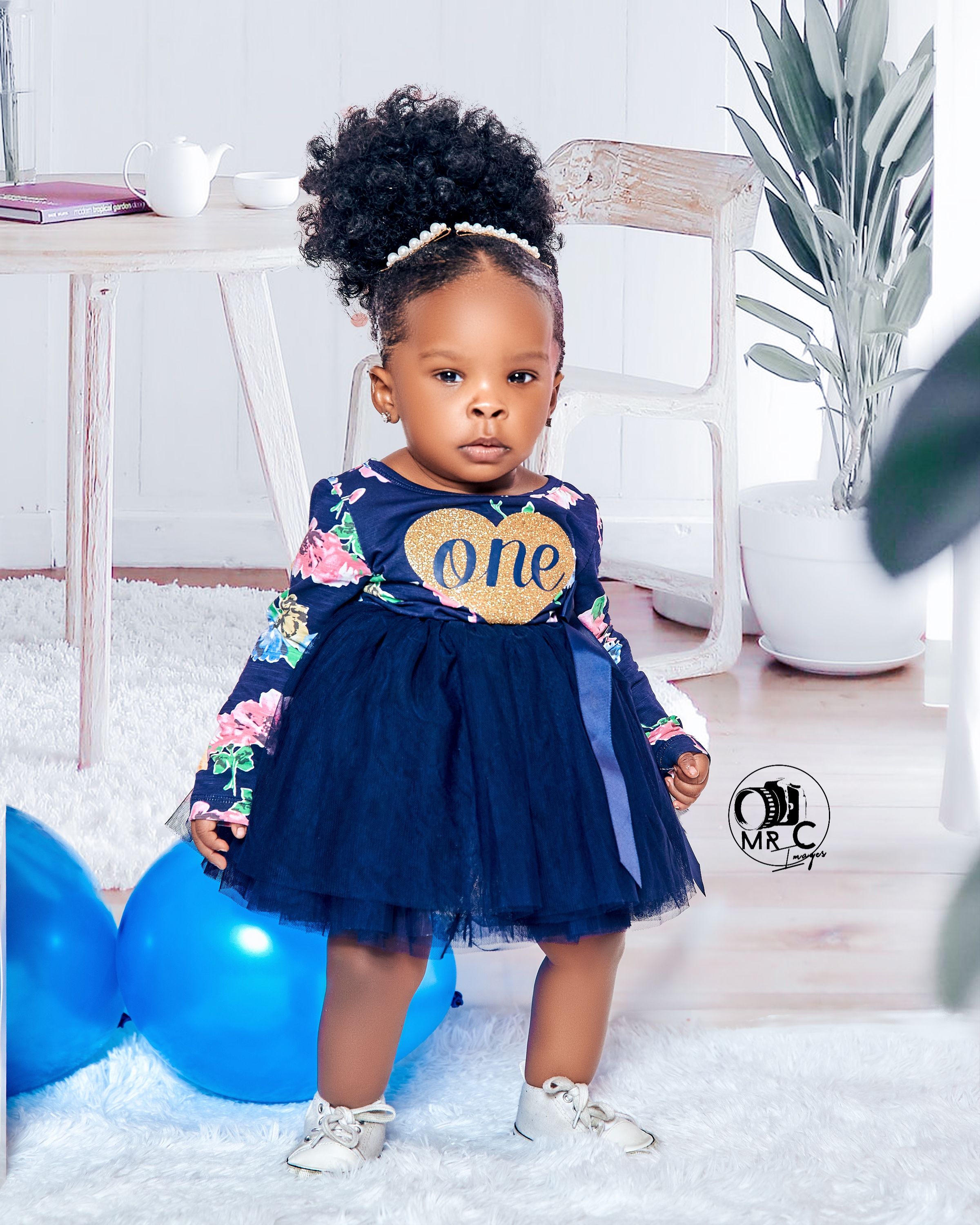 Child in navy blue tulle dress with 'one' emblem poses near white furniture and blue balloons.