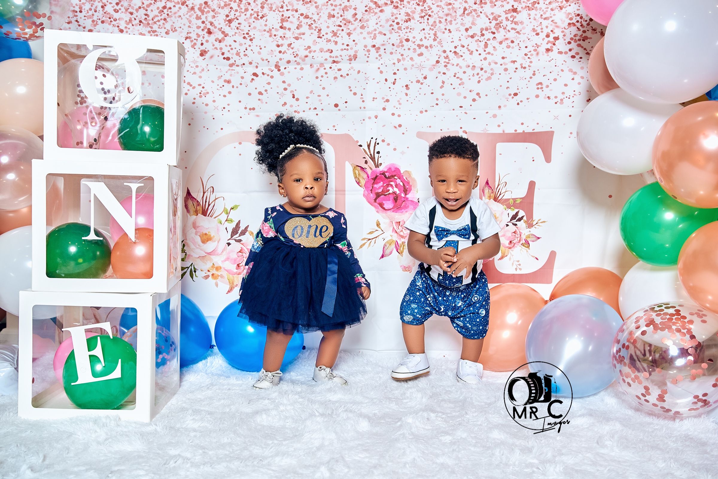 Children in festive outfits pose near decorative blocks spelling ONE with balloons and sparkles.