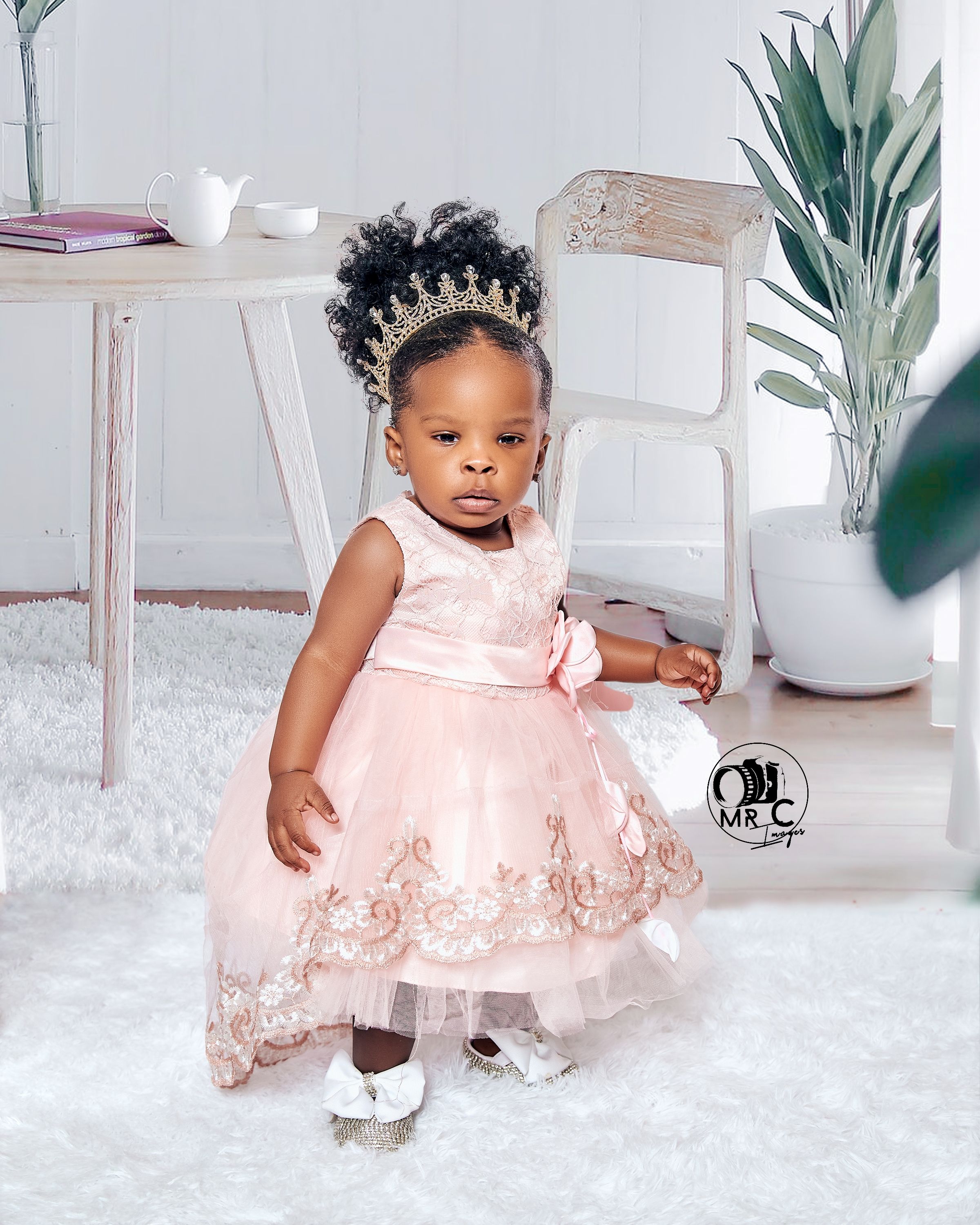 Small child in pink formal dress and crown posing near white furniture and indoor plants.