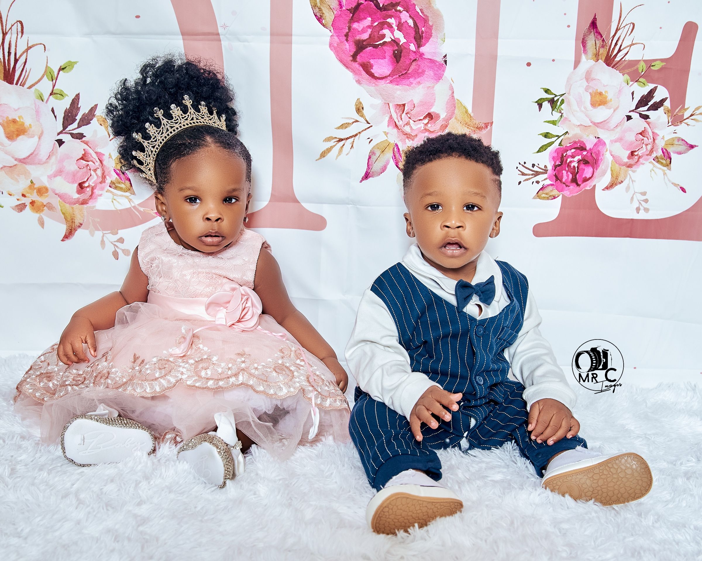 Two children in formal wear against floral backdrop, wearing pink dress and navy suit.