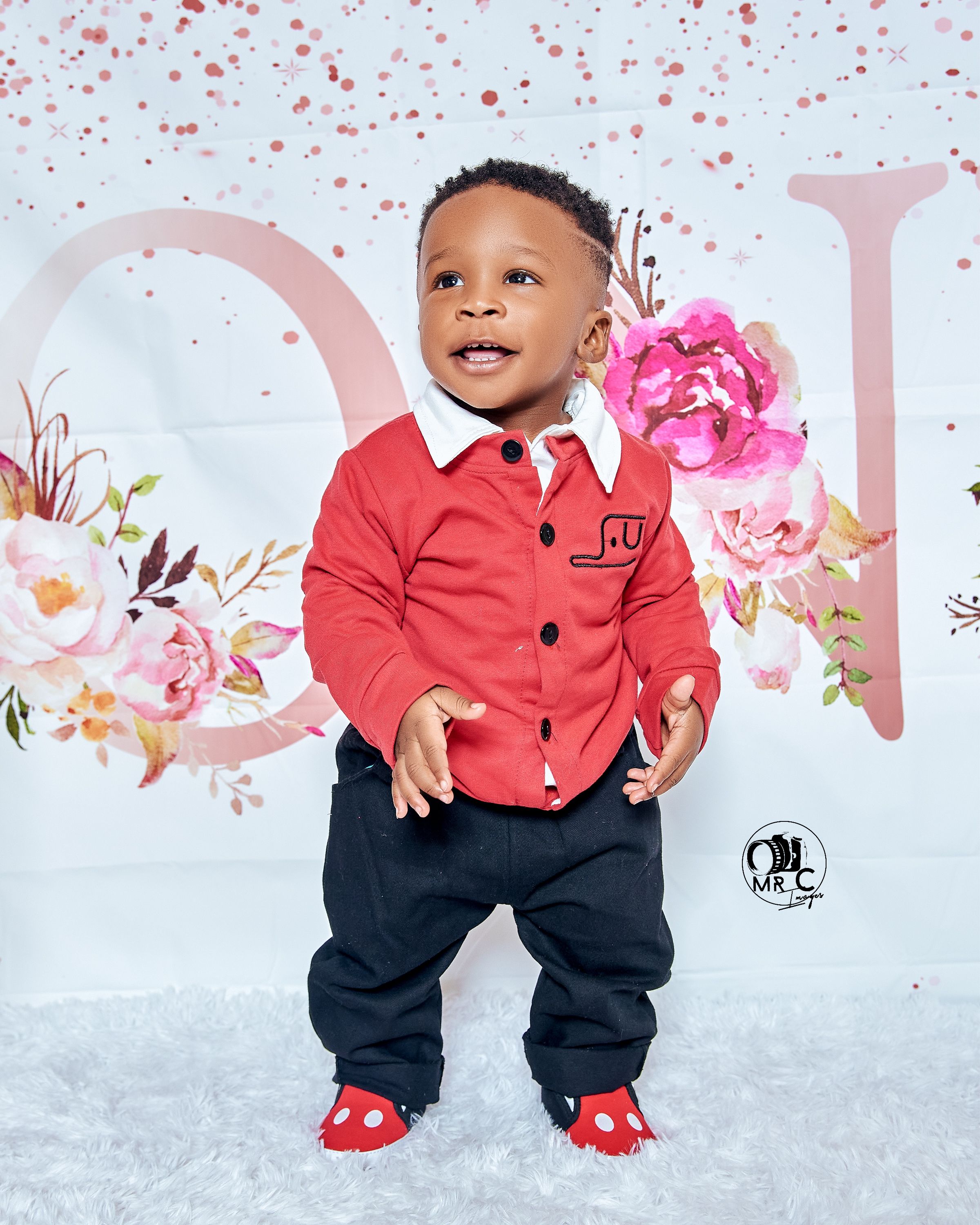 Toddler wearing red cardigan and navy pants against floral backdrop with pink decorations.