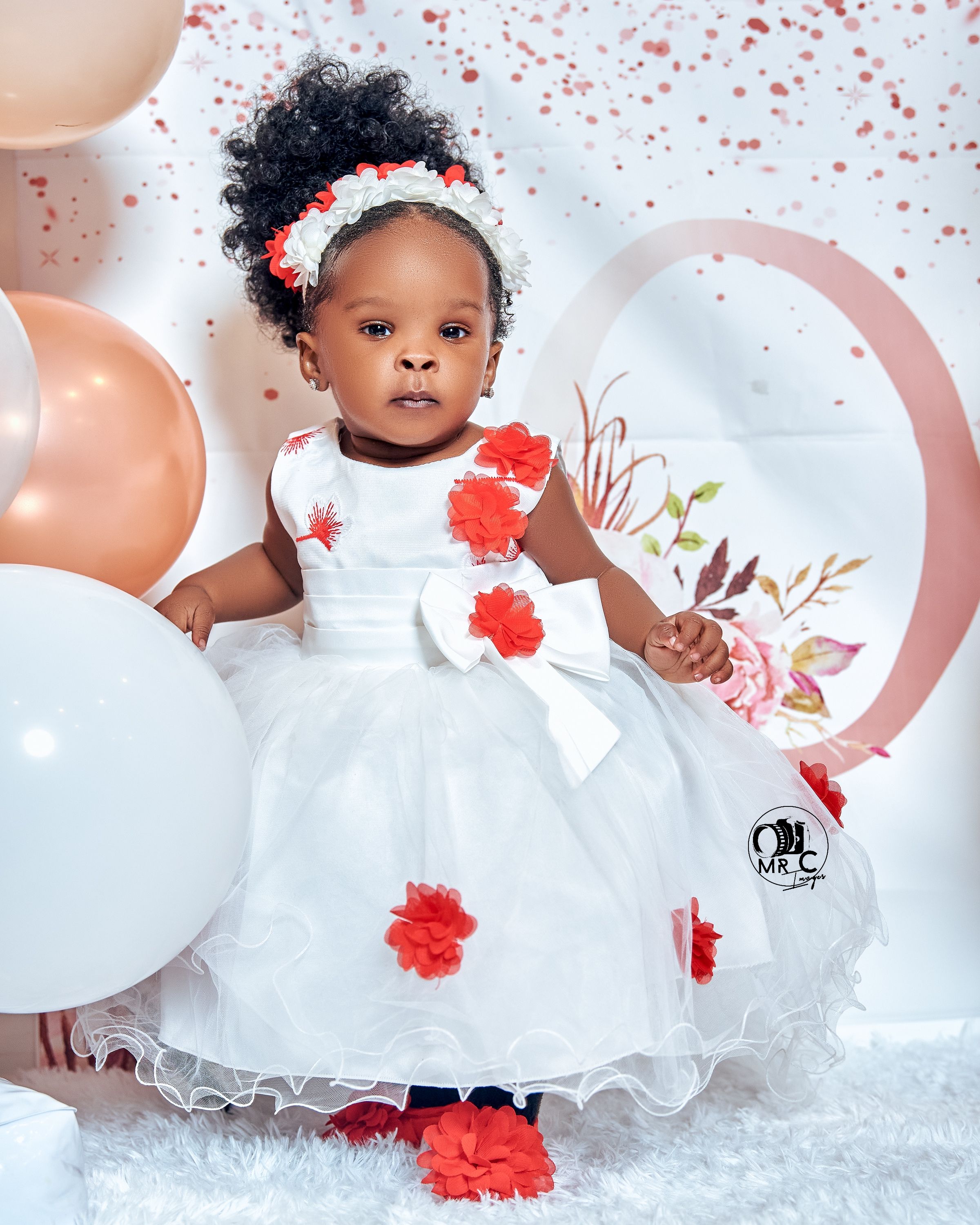 Toddler in white dress with red floral accents surrounded by balloons and pink decorative elements.