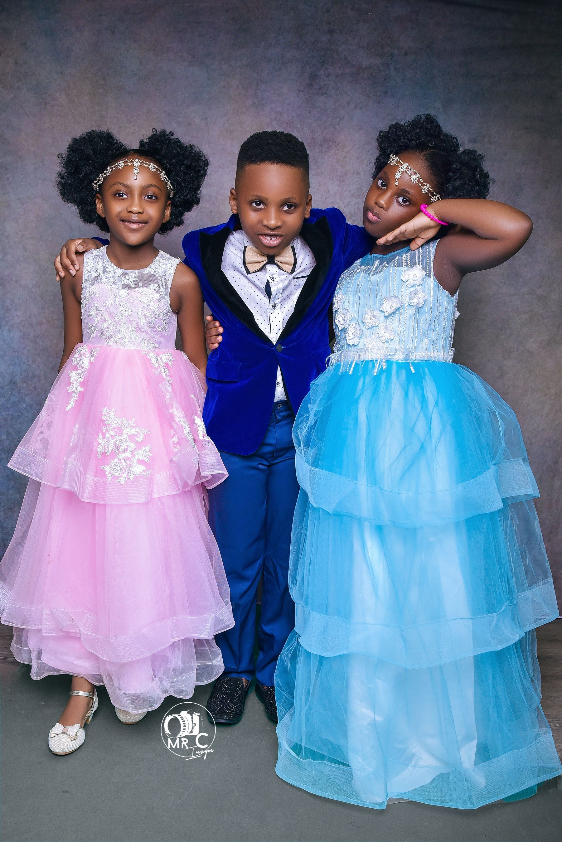 Three children in formal attire posing against grey backdrop, wearing pink, blue and navy outfits.