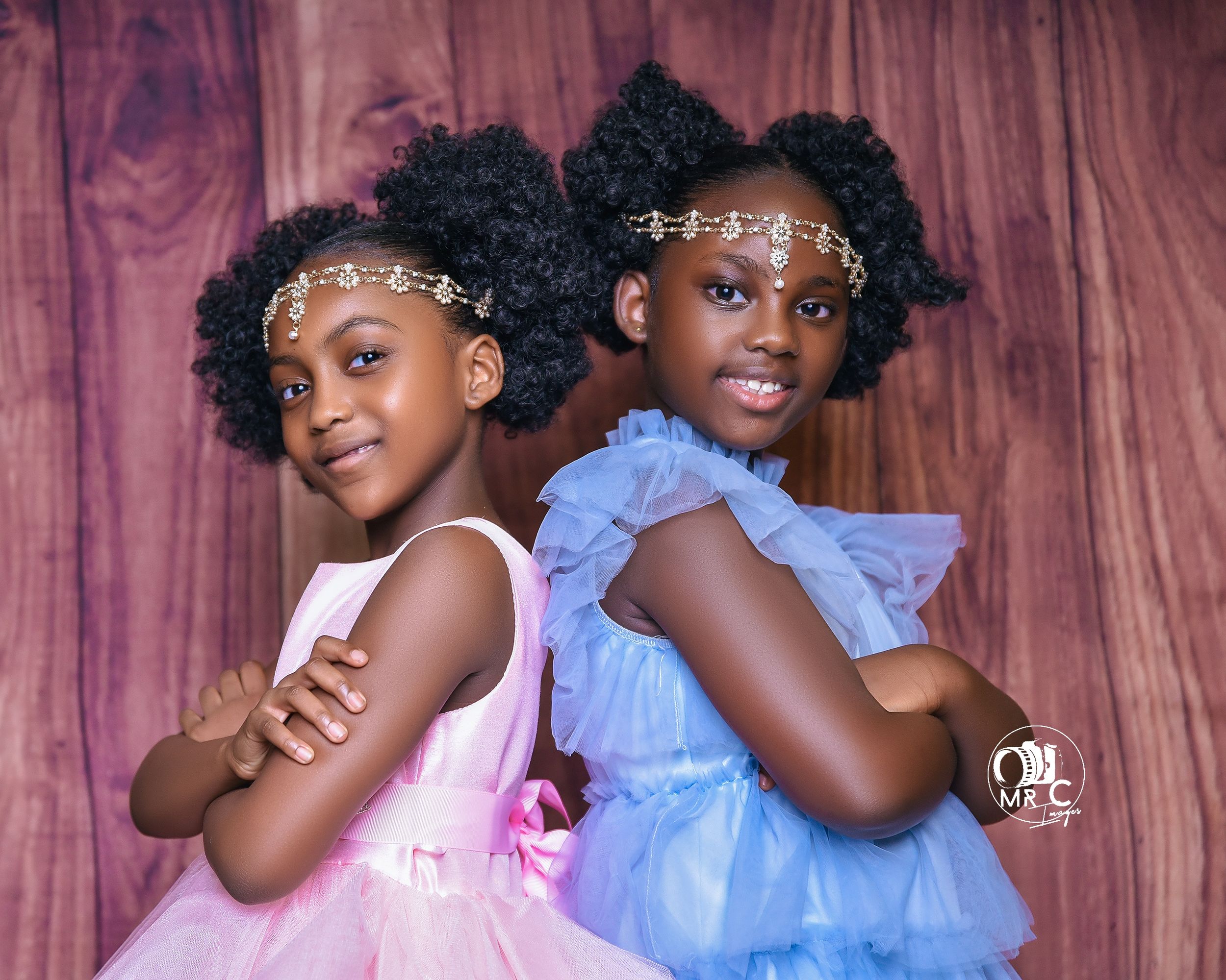 Two children in tulle dresses with sparkly headpieces pose back-to-back against a wooden backdrop.