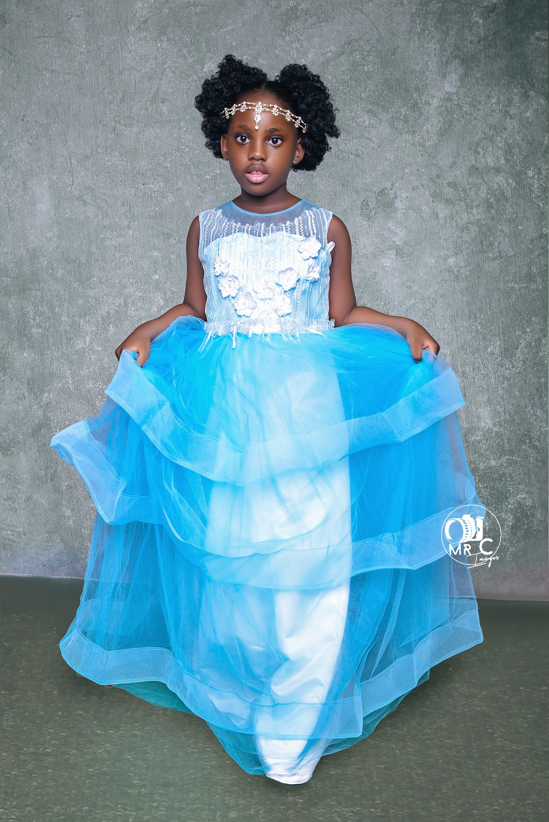 A young girl wearing a beautiful light blue and white princess ball gown with tulle layers poses against a gray background.