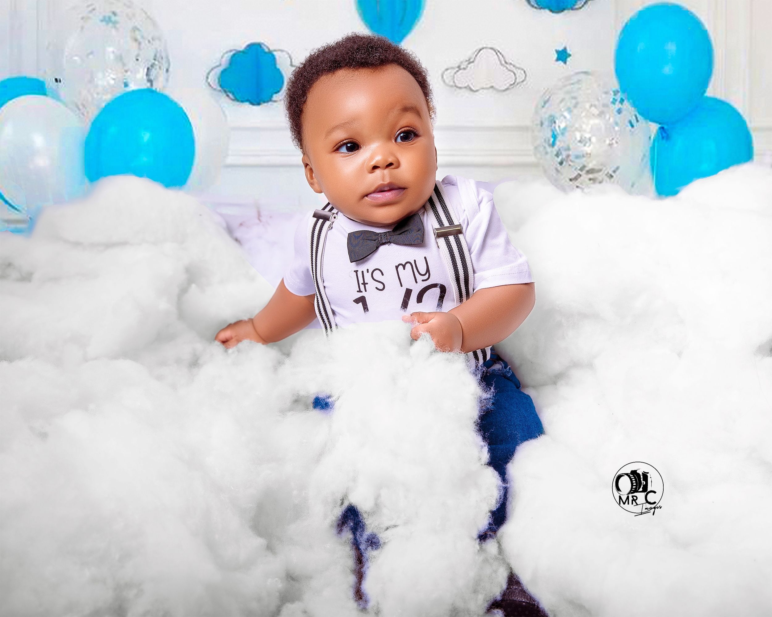 Toddler in casual attire surrounded by blue and white balloons with cloud-like decorations.