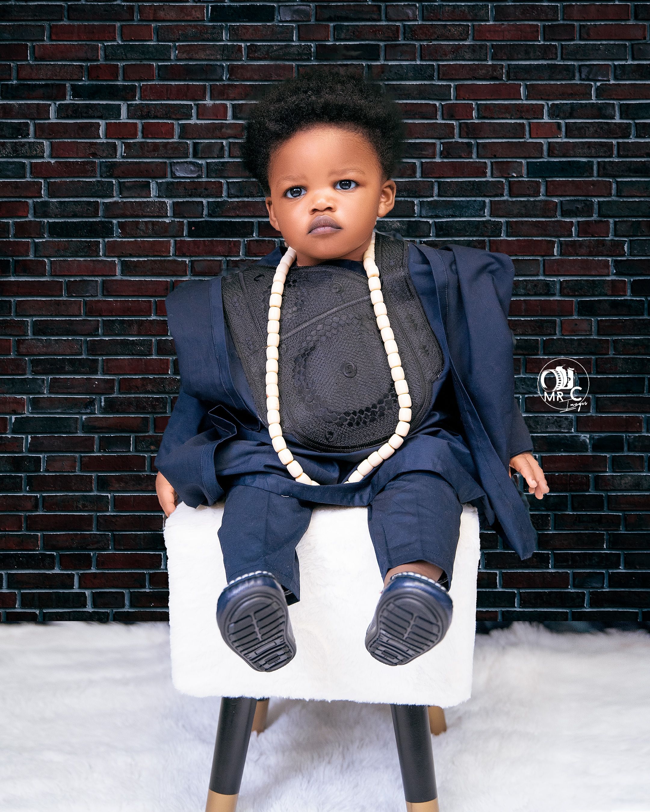 Stylishly dressed toddler in navy suit with white rope accessory seated on modern white chair against brick wall.
